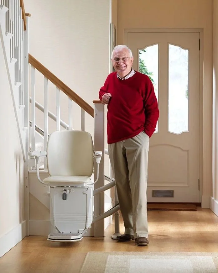 A smiling man stands in a brightly lit home hallway next to a white stairlift installed at the base of a wooden staircase.