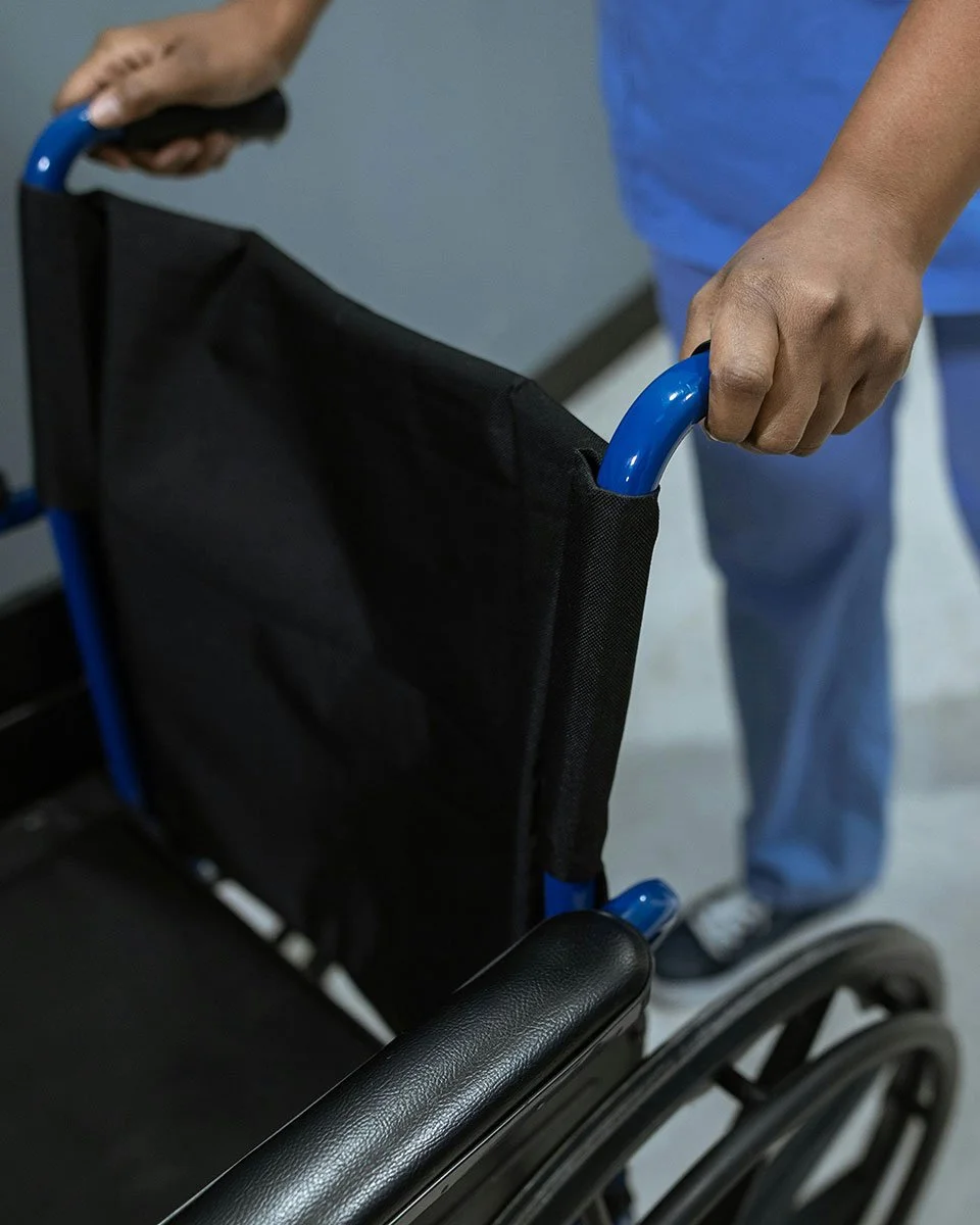 A close-up view of a healthcare worker in blue scrubs gripping the blue handles of a black manual wheelchair to provide mobility assistance.