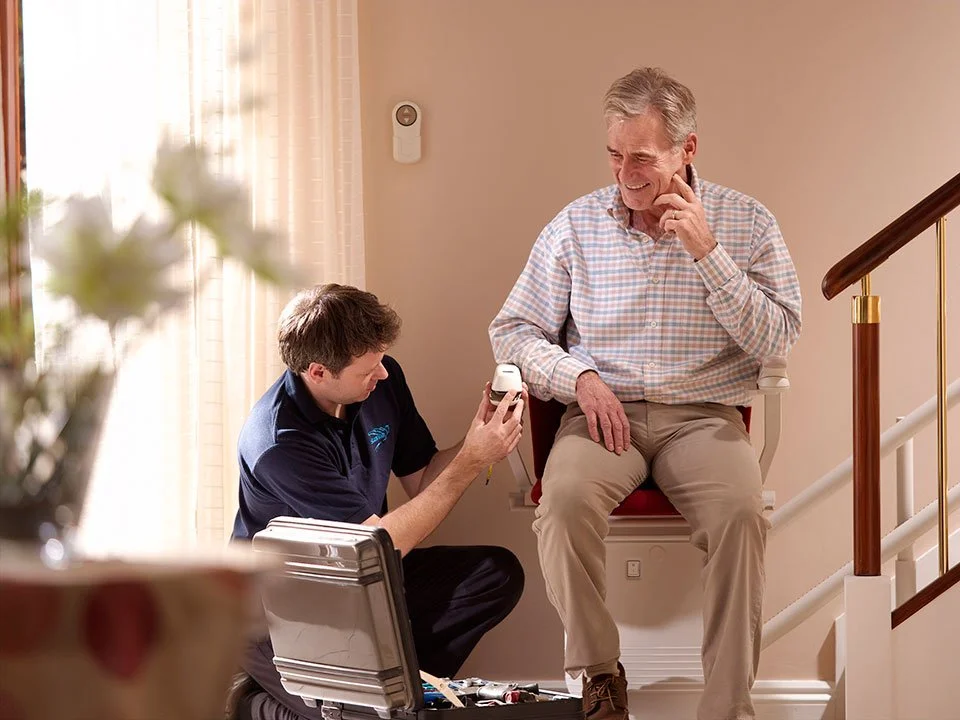 A service technician kneels beside a man seated on a stairlift to demonstrate a hand-held remote control while his tool kit sits open on the floor.