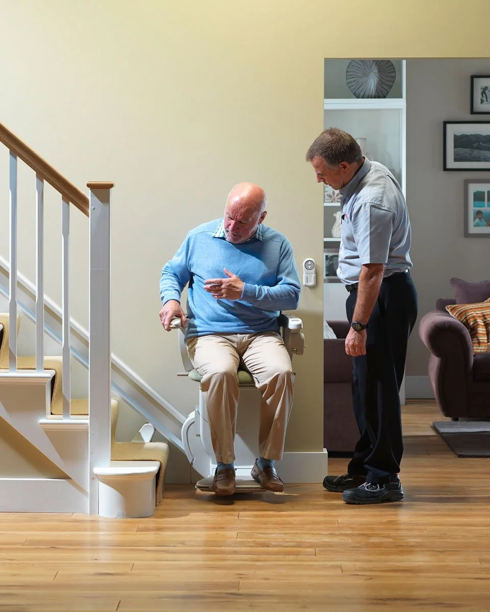 An older man sits on a stairlift while a technician provides a demonstration of the controls at the bottom of a home staircase.