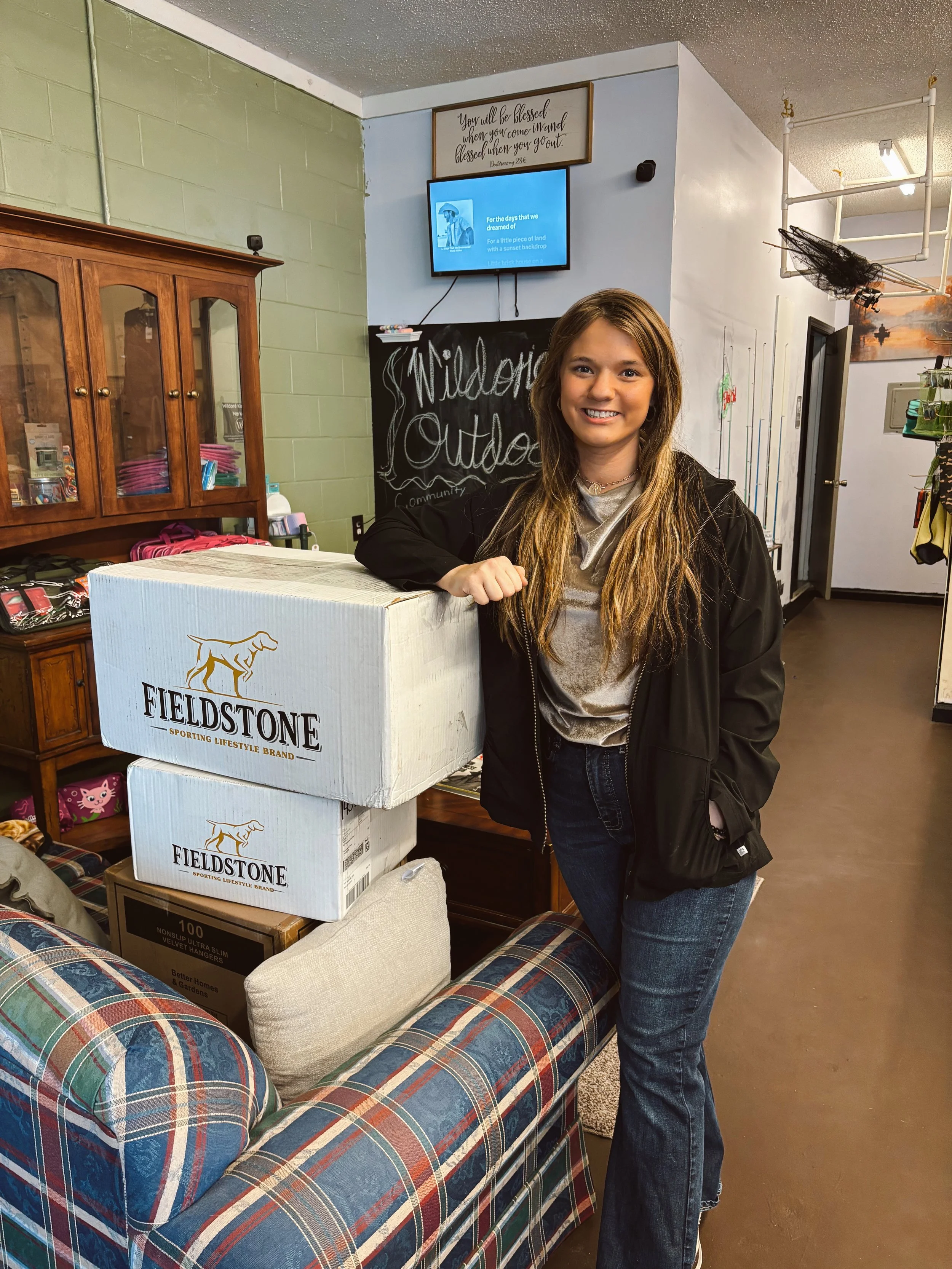 Young woman with long blonde hair smiling, standing next to boxes of Fieldstone brand dog food in an indoor setting with a plaid couch nearby.