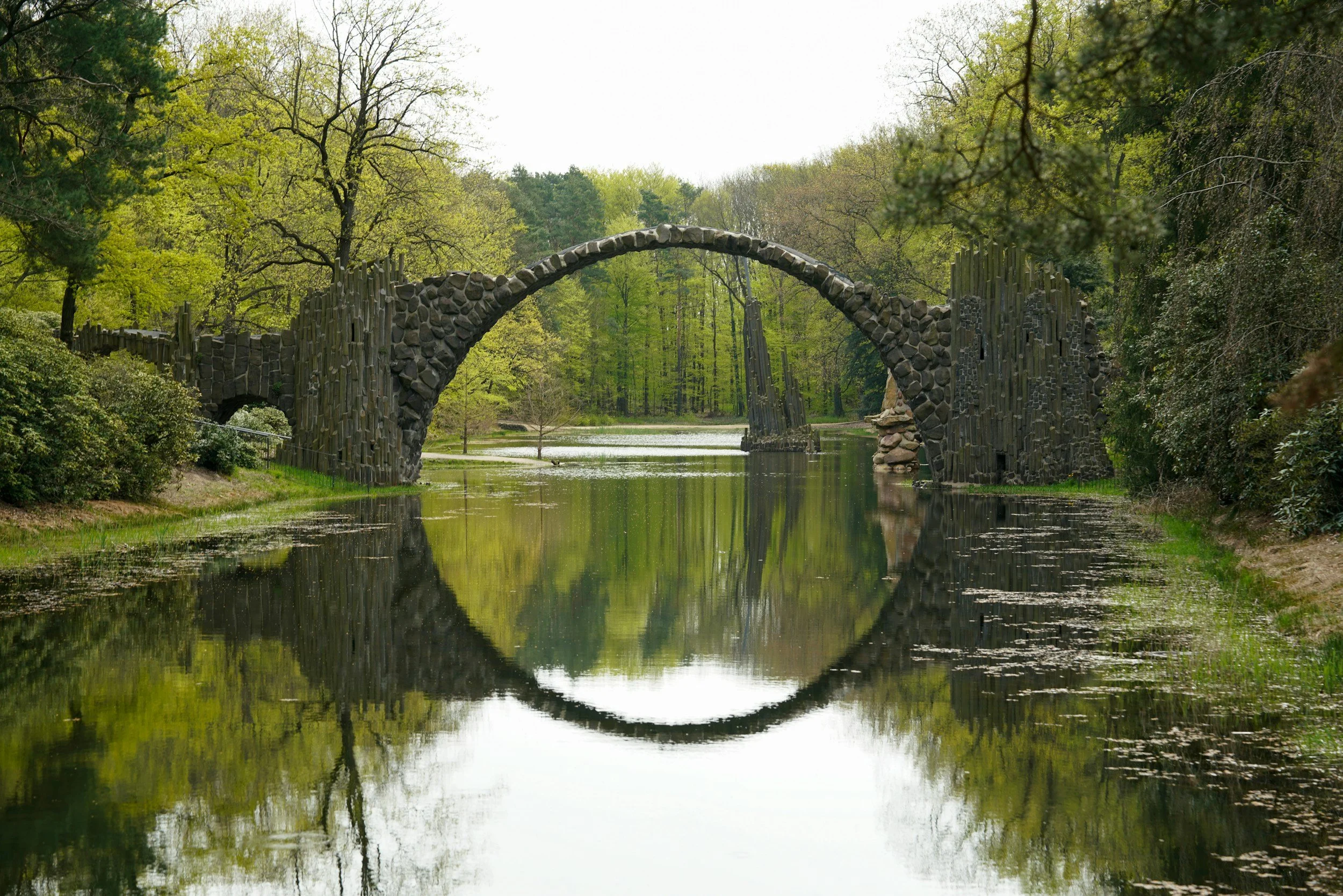 A stone arch bridge reflecting in a calm body of water surrounded by green trees.