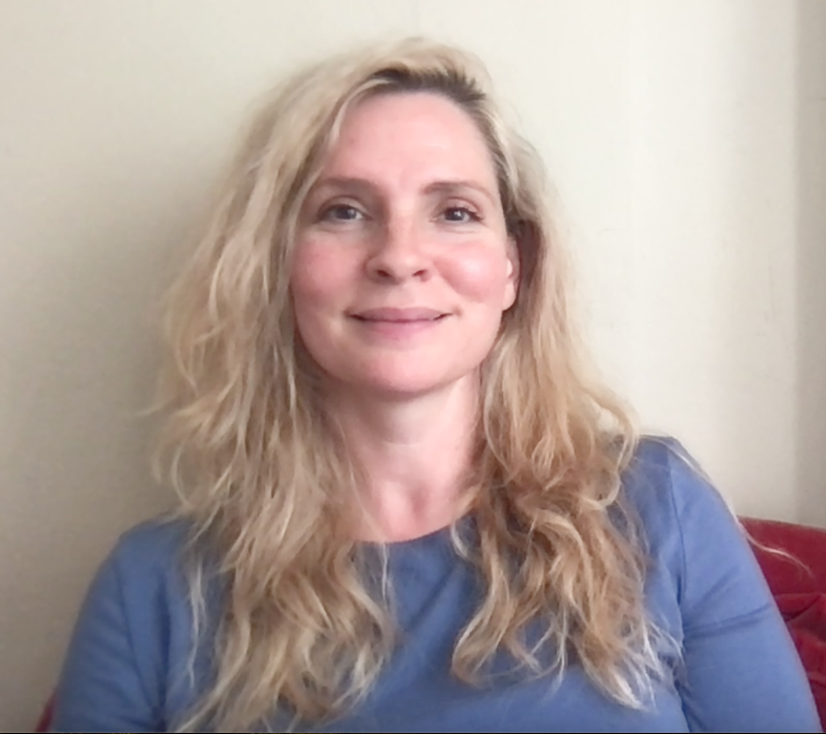 A woman with long, wavy blonde hair and a light complexion, smiling at the camera against a plain, cream-colored wall.