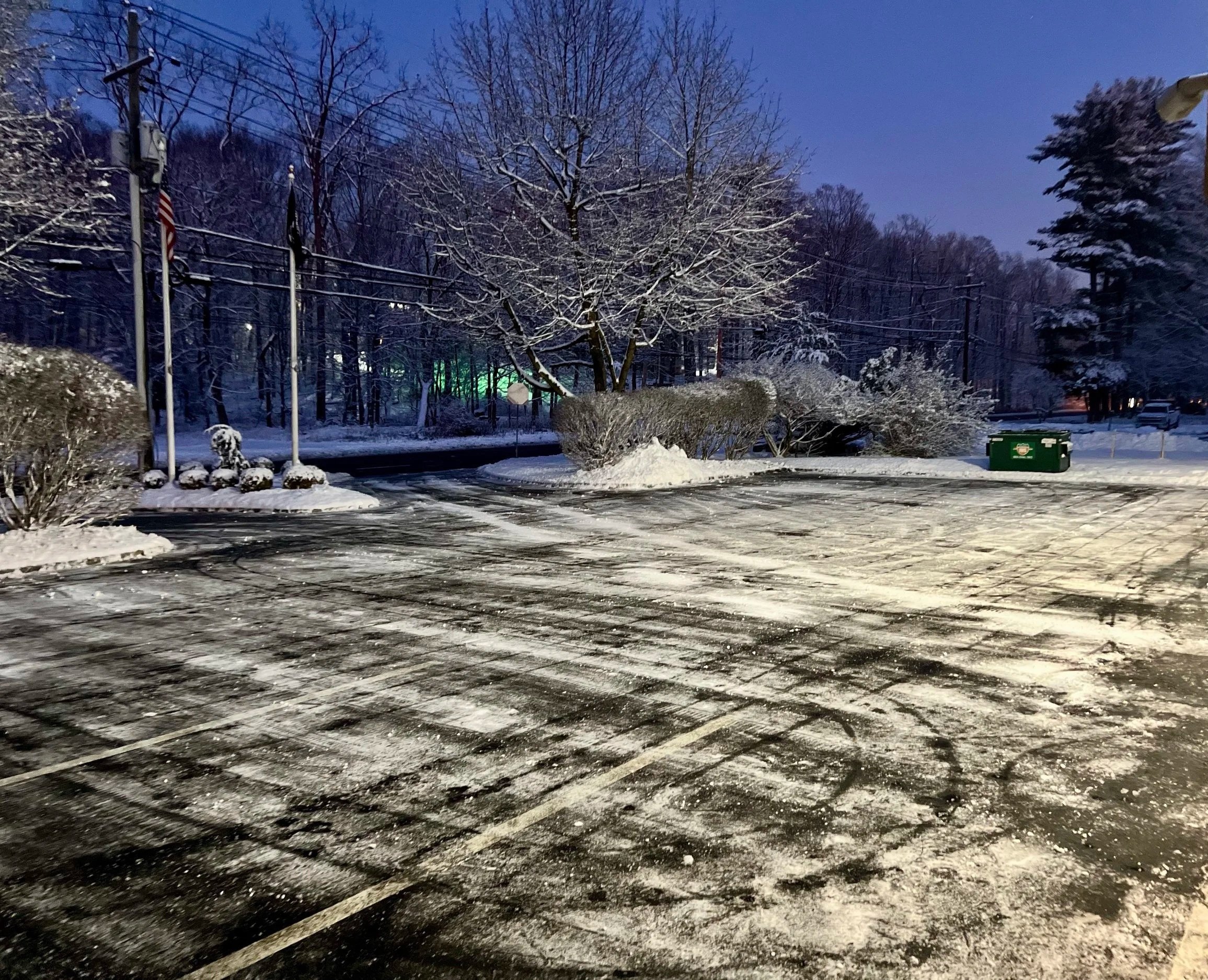 Snow-covered parking lot at dusk with trees, bushes, flags, a green trash bin, and tire tracks on the icy surface.