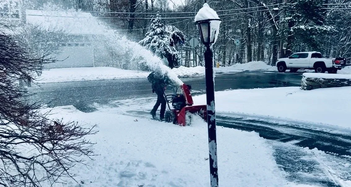 Person operating a snowblower on a snow-covered sidewalk during a snowstorm in a residential neighborhood.