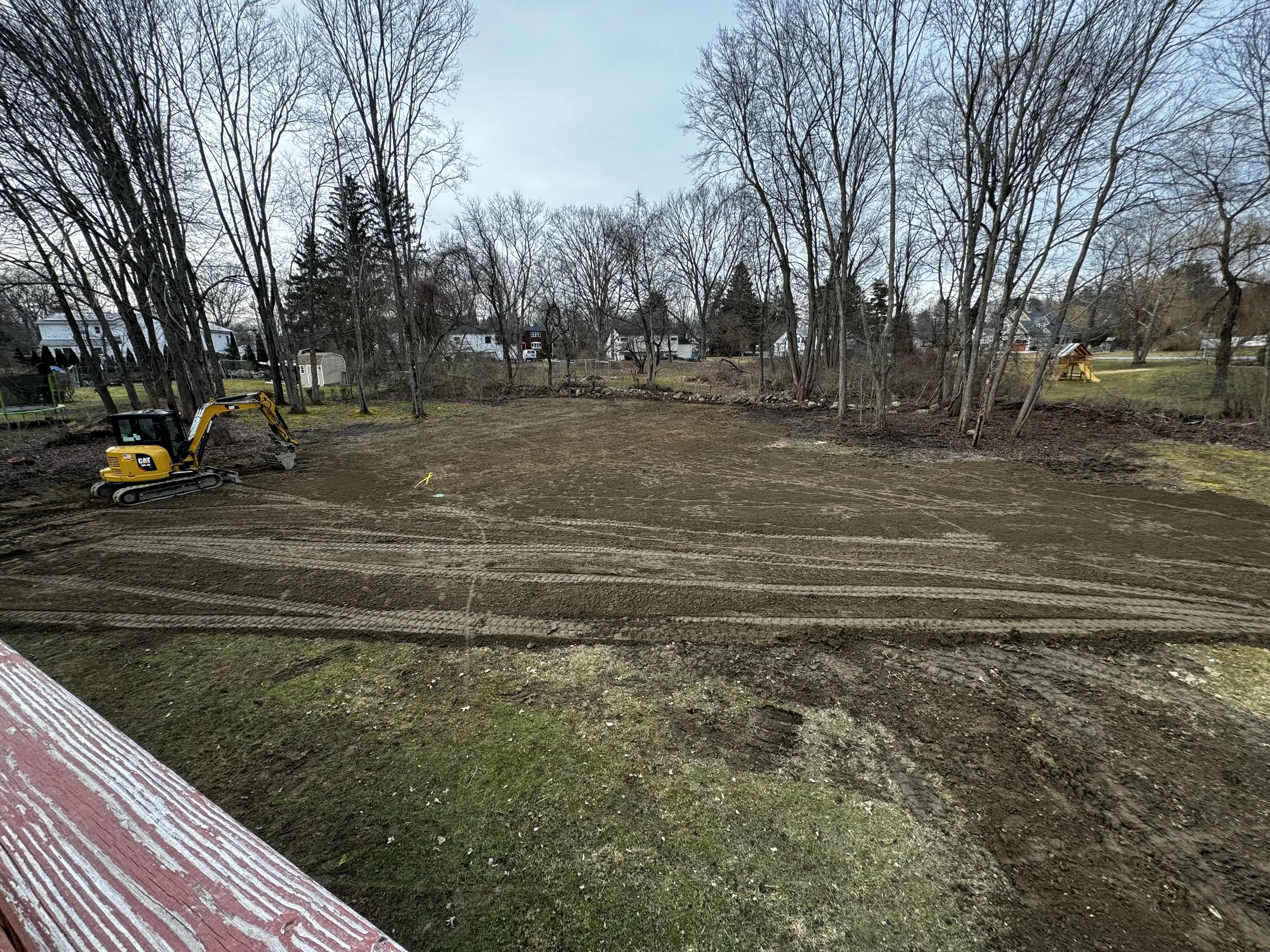A backyard being prepared for construction with a small yellow excavator parked on dirt, surrounded by leafless trees and residential houses in the background.