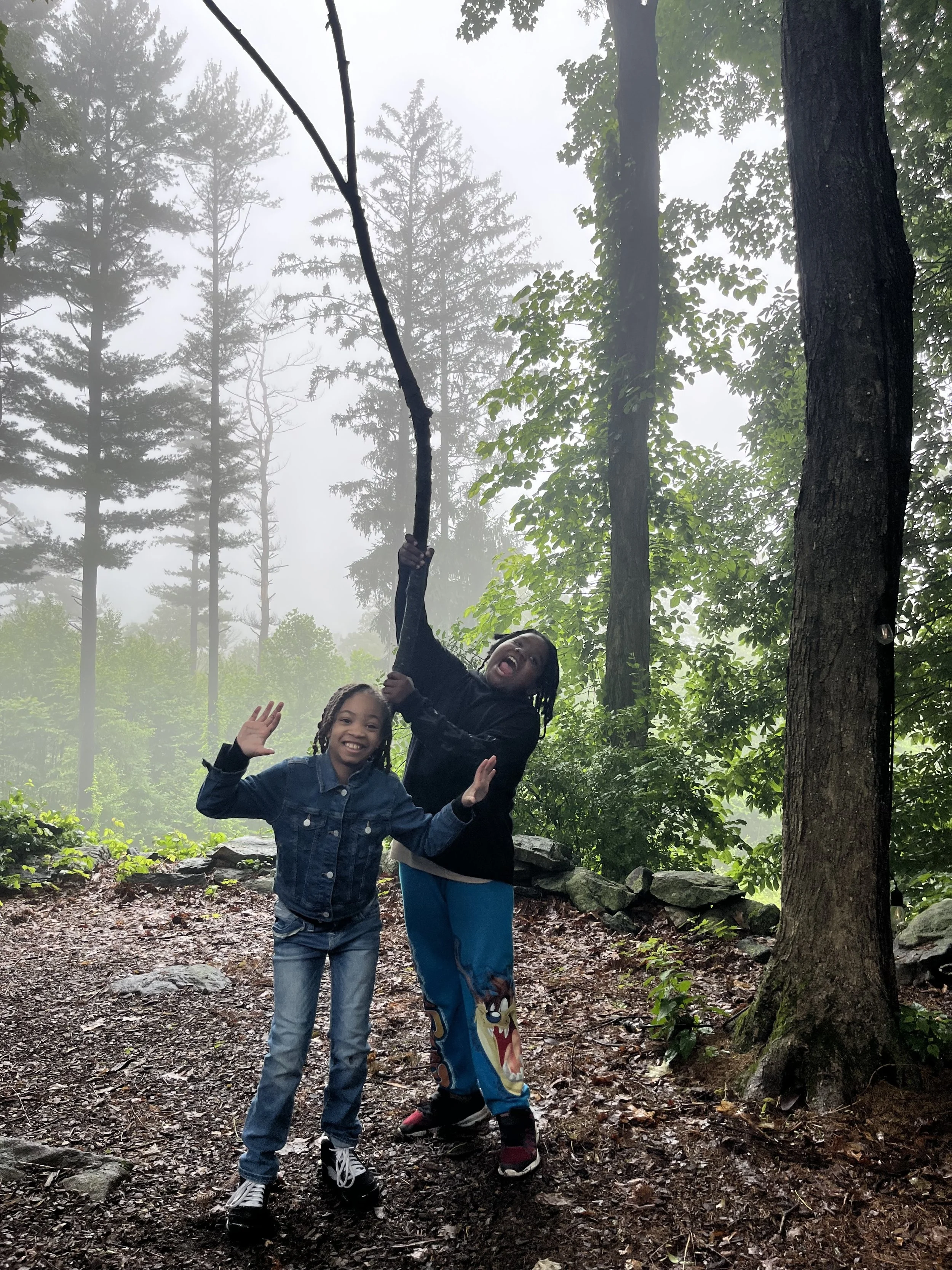 Two children, a girl and a boy, standing on a muddy forest trail with tall trees and fog in the background. The girl is smiling and waving at the camera, while the boy is holding onto a tree branch with both hands, looking excited.