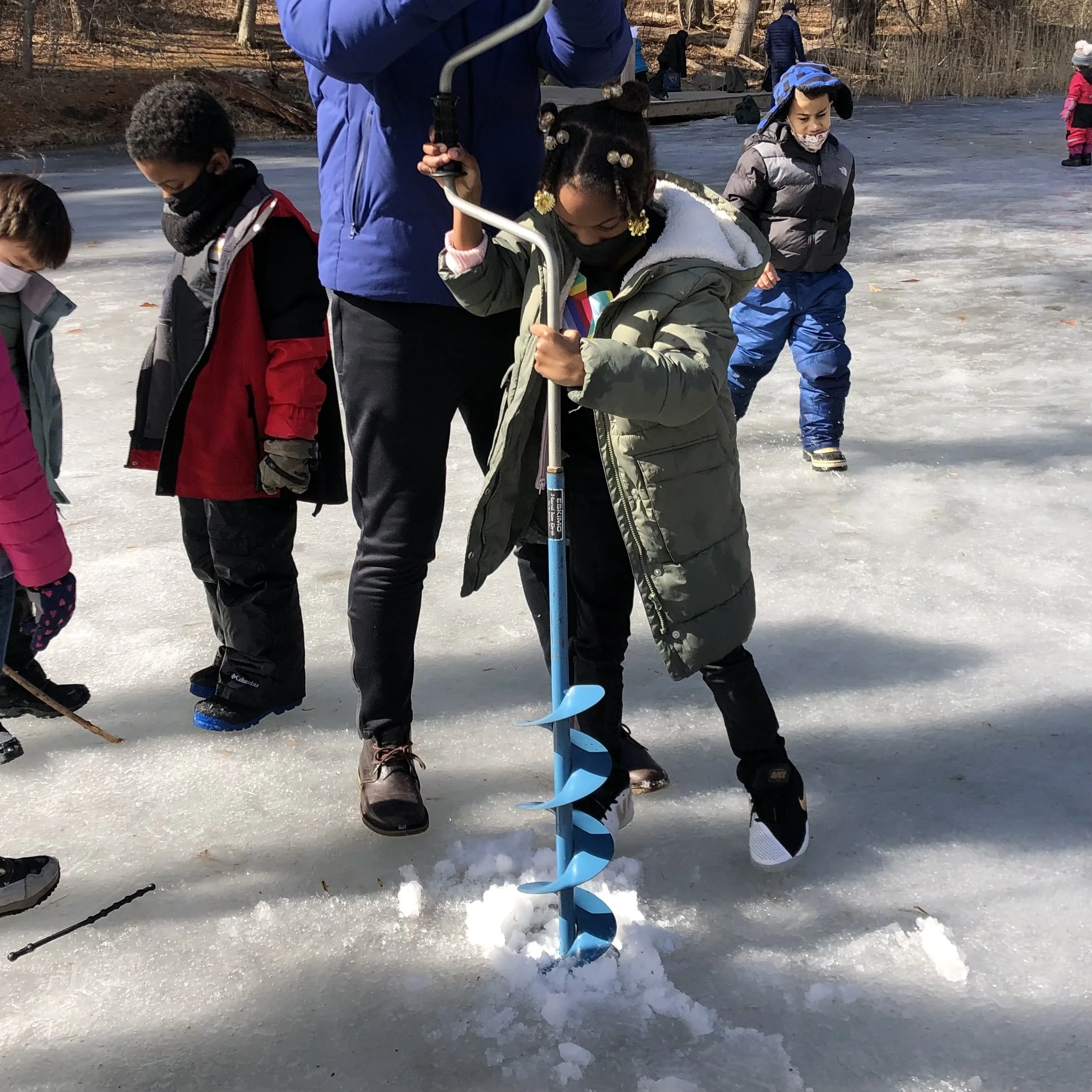 A group of children ice fishing on a frozen pond with an adult holding a blue ice auger. The children are dressed in winter clothing and are standing on ice, some watching while one girl is operating the ice auger to drill a hole.