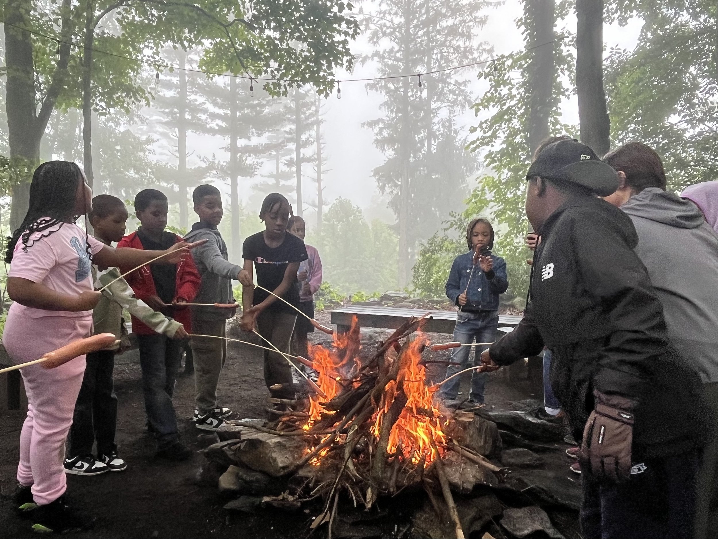 City to Sanctuary students and teachers gathered around a campfire in a foggy, wooded outdoor setting, roasting food on sticks.