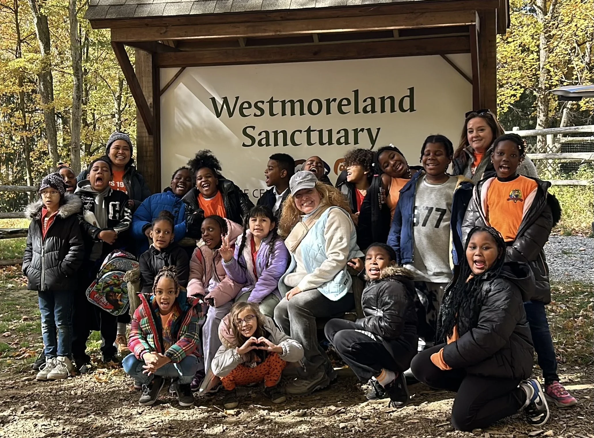 A group of children and adults smiling and posing for a photo in front of a sign that reads "Westmoreland Sanctuary" in a forested area during fall.