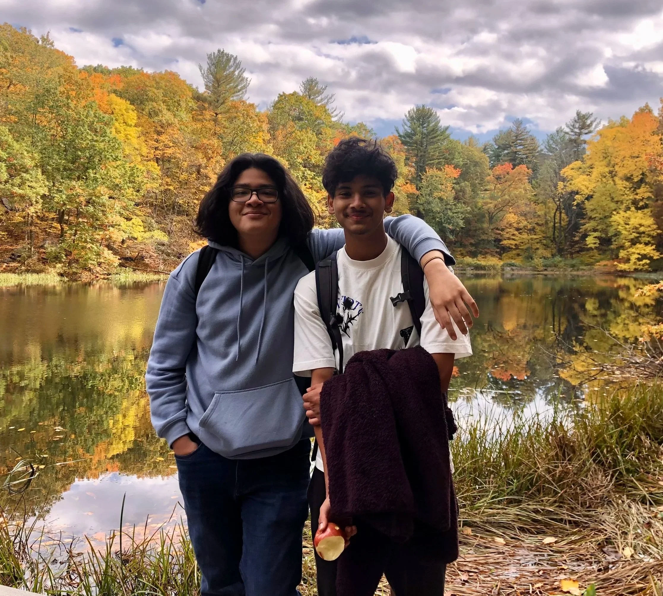 Two young people standing close together near a lake with autumn trees in the background, one with their arm around the other's shoulder, smiling at the camera.