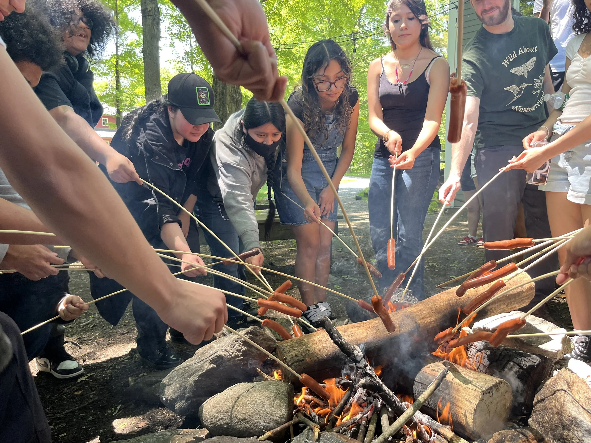 Group of people roasting hot dogs over an outdoor campfire in a wooded area.