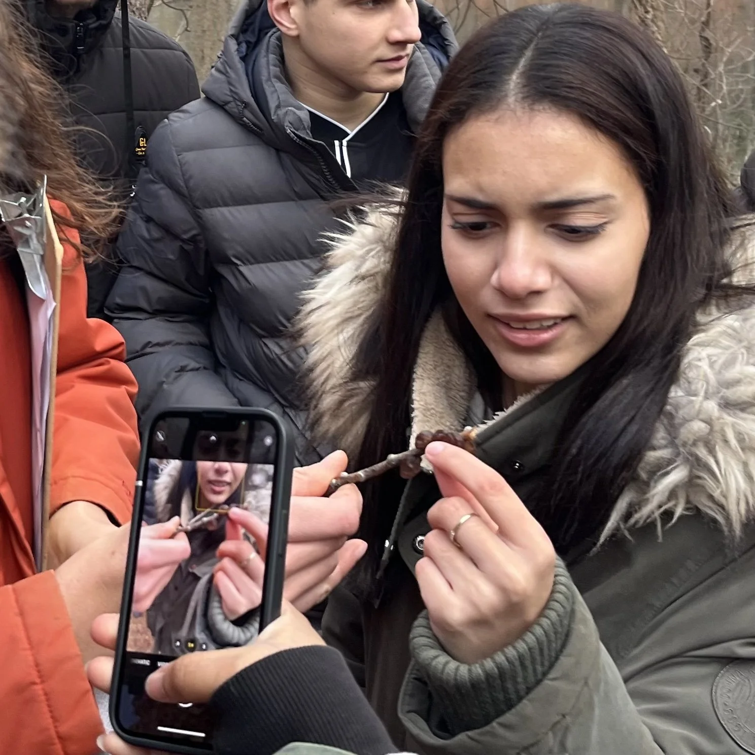 A woman with long dark hair and a fur-lined jacket holds a small stick with what a small insect attached to it, during a City to Sanctuary program.