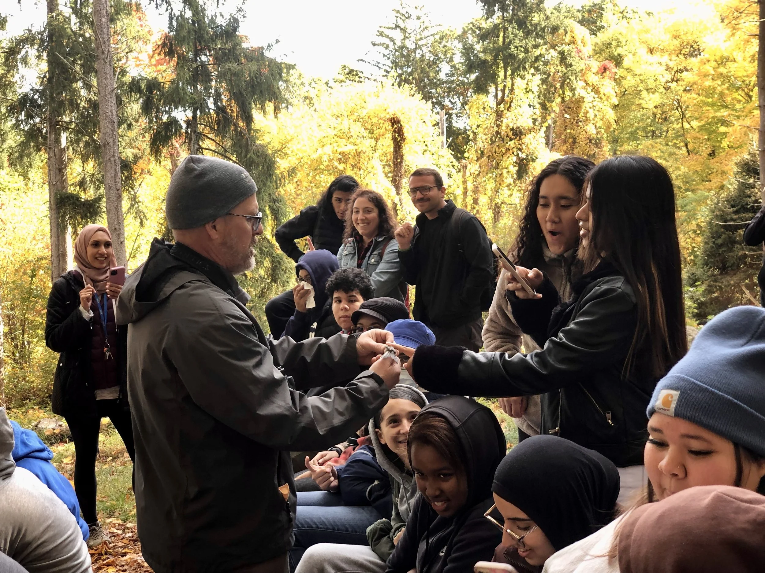 A group of young people, including children and adults of diverse ethnicities, gathered outdoors in a forest with colorful autumn trees. An older man in a gray beanie and glasses appears to be signing an item for a girl, while others are smiling, taking photos, and engaging with each other.