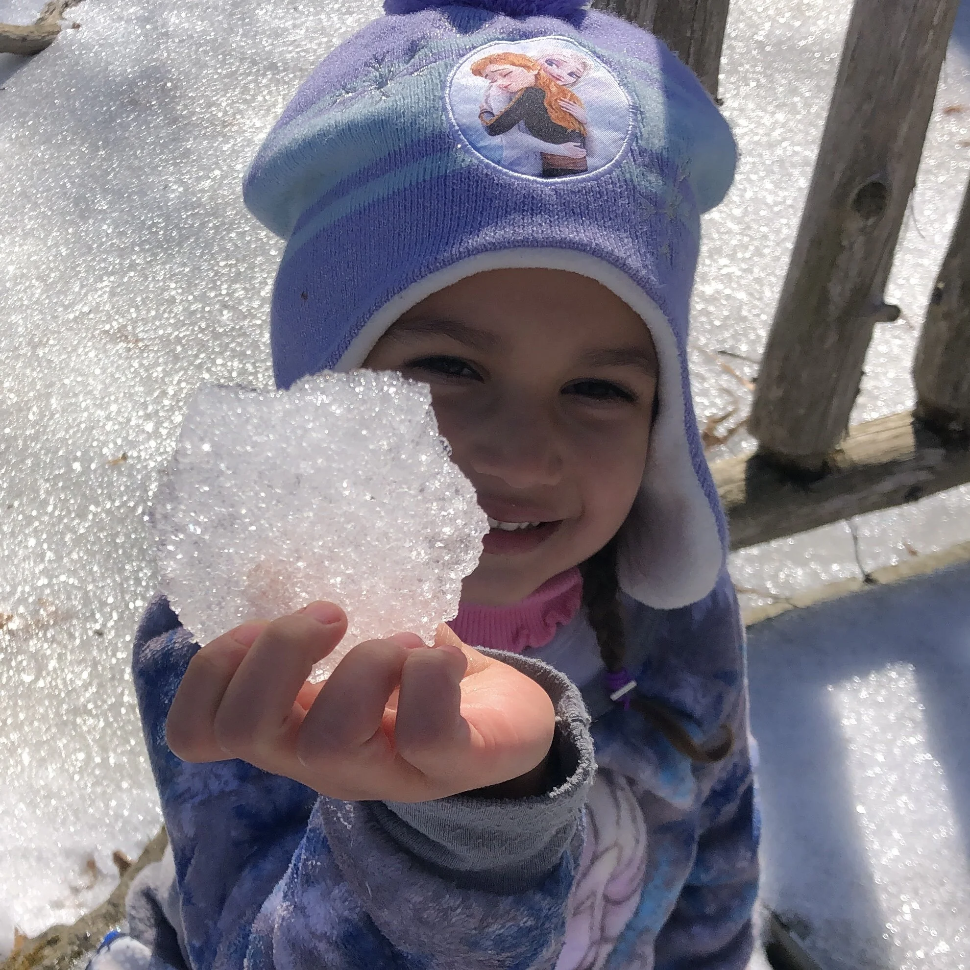 A young girl holding a chunk of snow, smiling, outdoors in bright sunlight, wearing a purple hat with a cartoon character, and a colorful jacket, near a wooden fence.