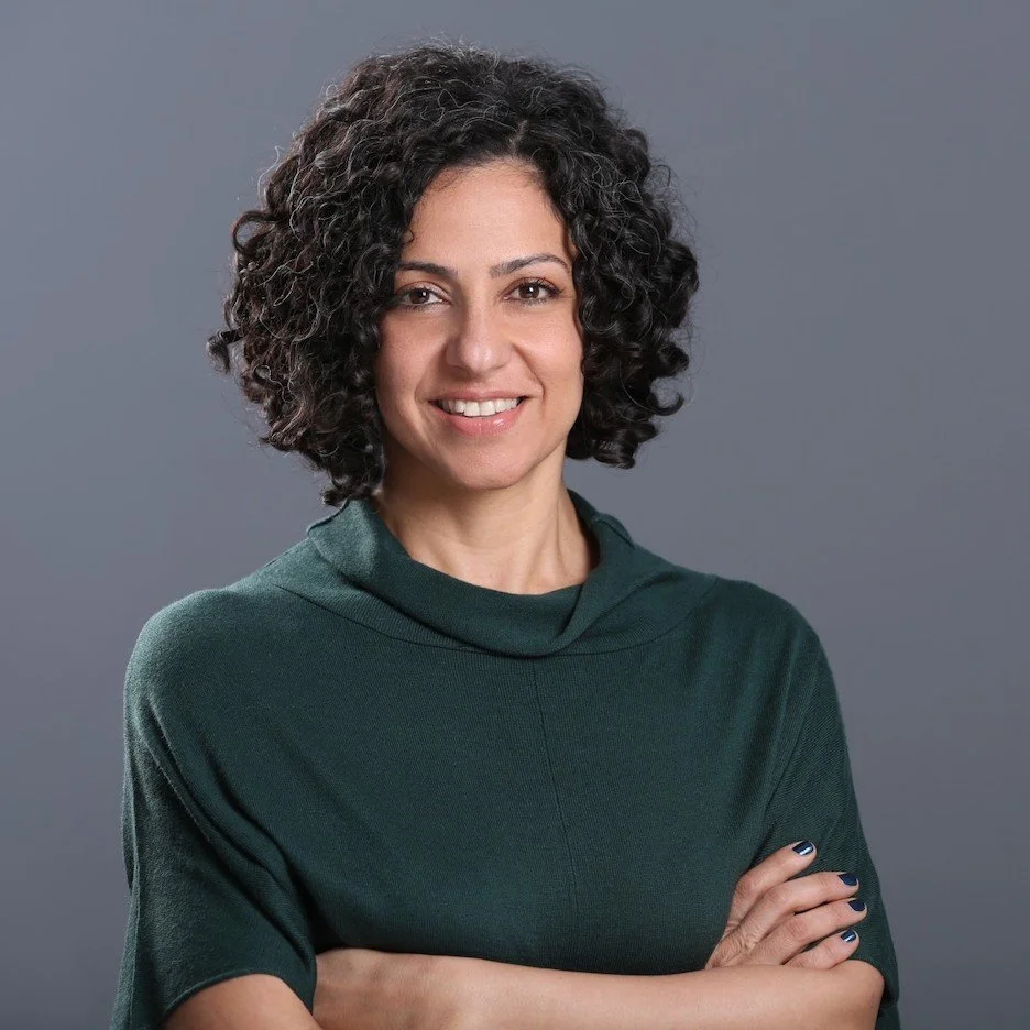 A woman with curly dark hair smiling, wearing a dark green top, standing against a gray background.
