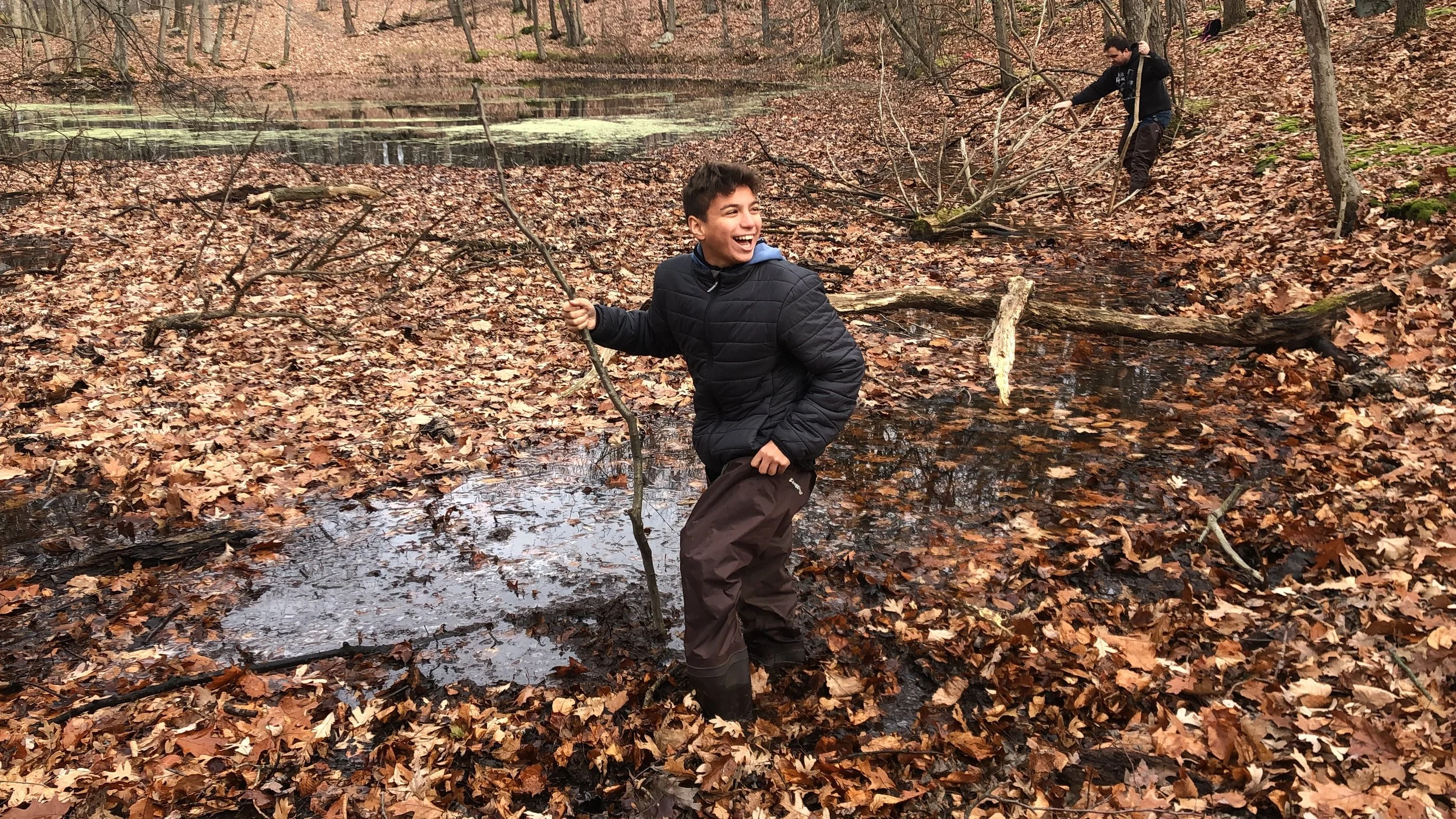Boy smiling and holding a stick in a flooded, leaf-covered forest during the City to Sanctuary nature-based learning program.