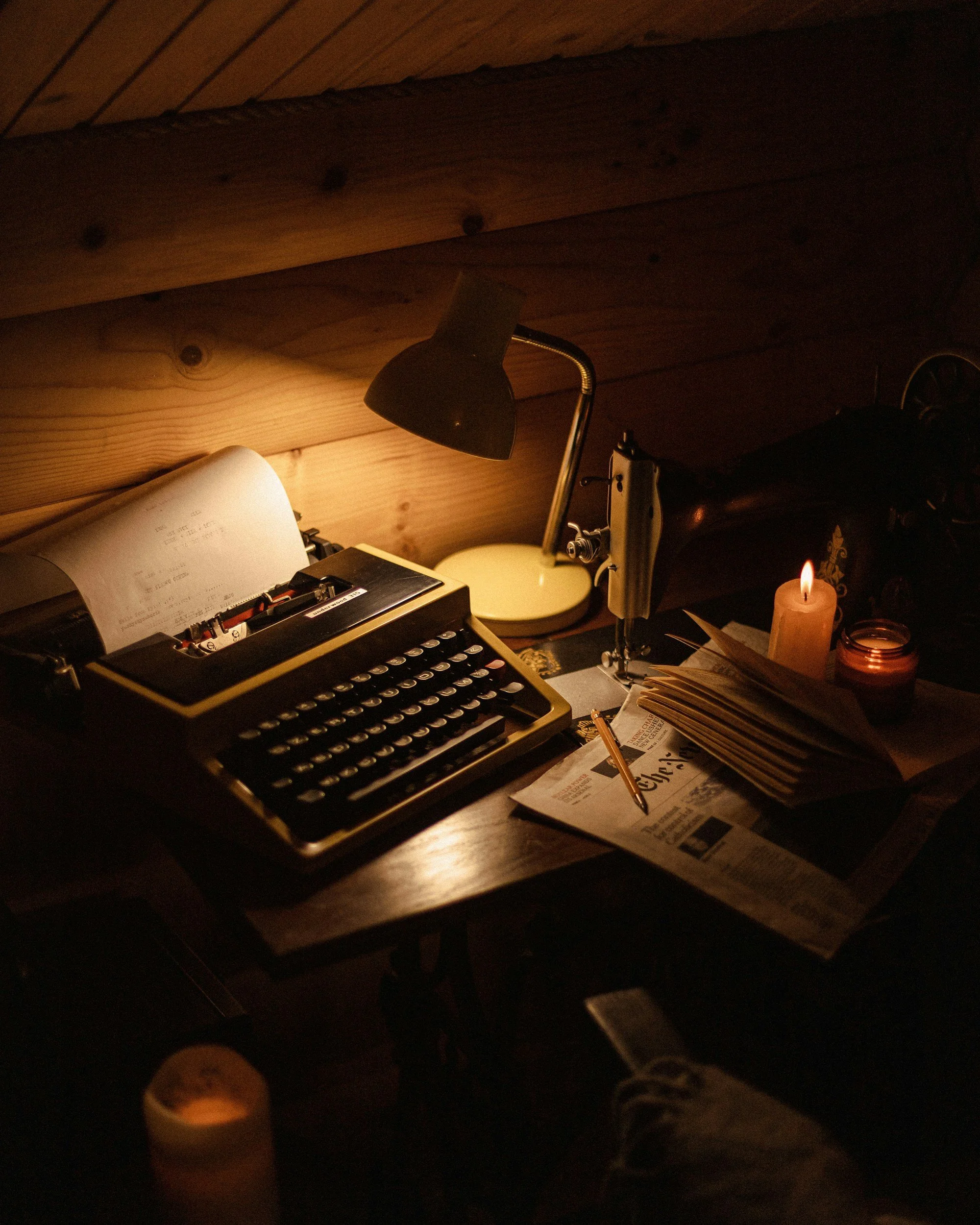 A dimly lit wooden desk with a vintage typewriter, a desk lamp, an open book, a candle, and a newspaper.
