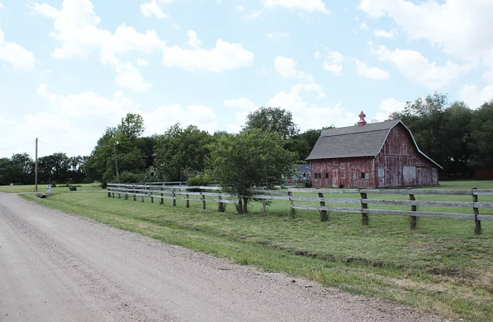 A rural scene with a gravel road, a rustic red barn with peeling paint, a green tree, and a wooden fence under a partly cloudy sky.