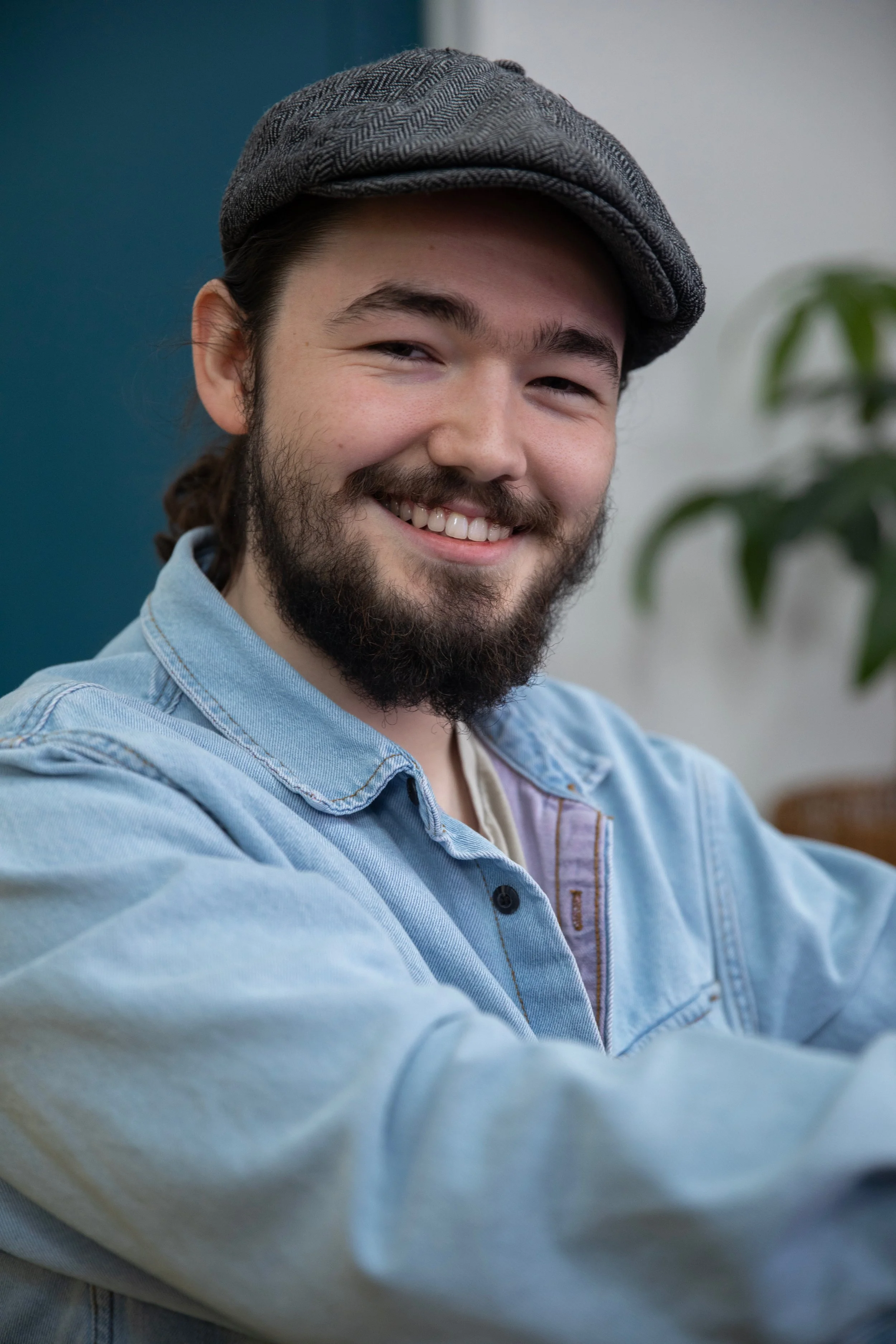 A young man with a beard and mustache smiling, wearing a gray cap and a light blue denim jacket, in an indoor setting with a plant in the background.