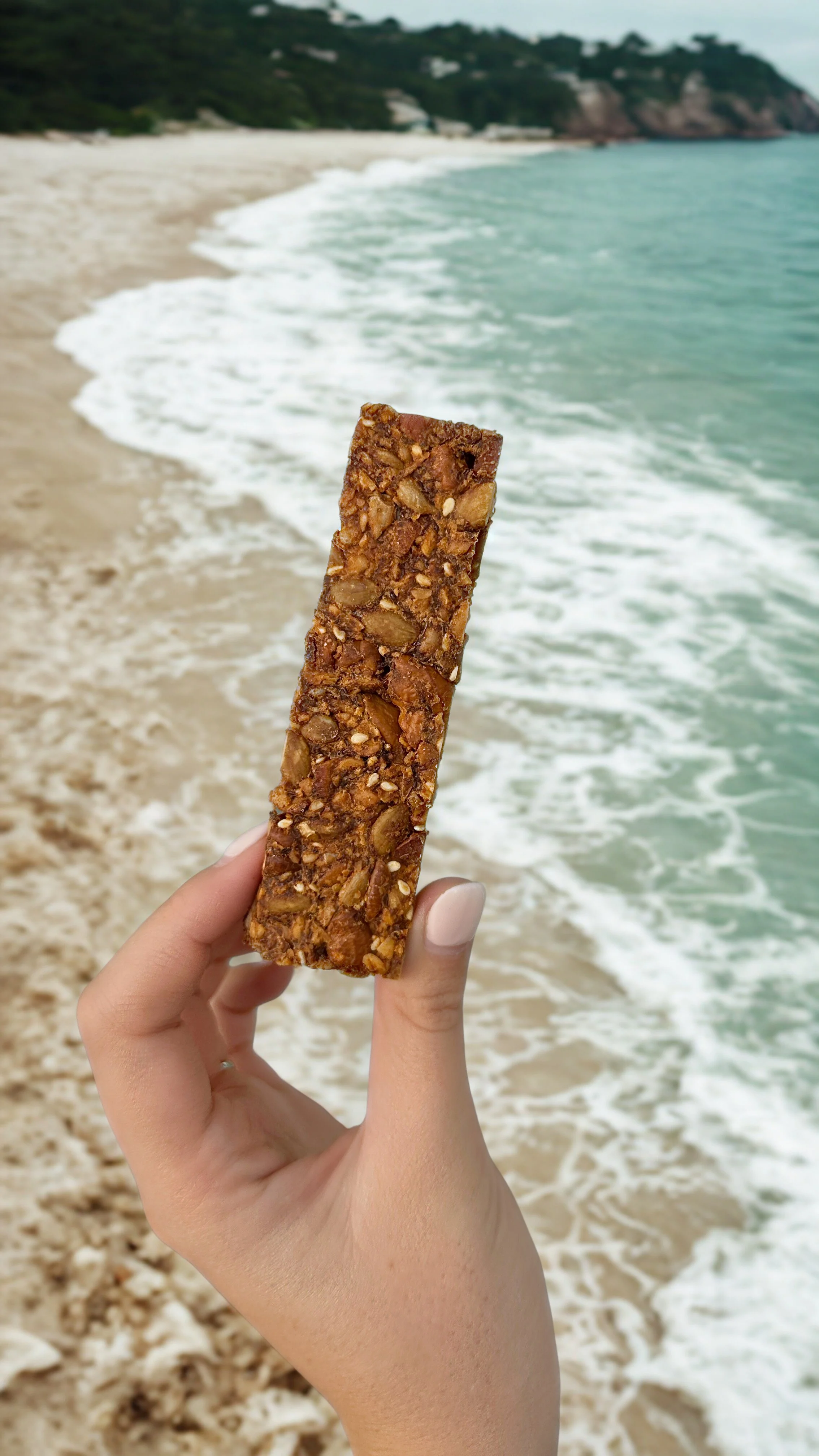 hand holding a noci granola bar with an ocean and sandy beach in the background.