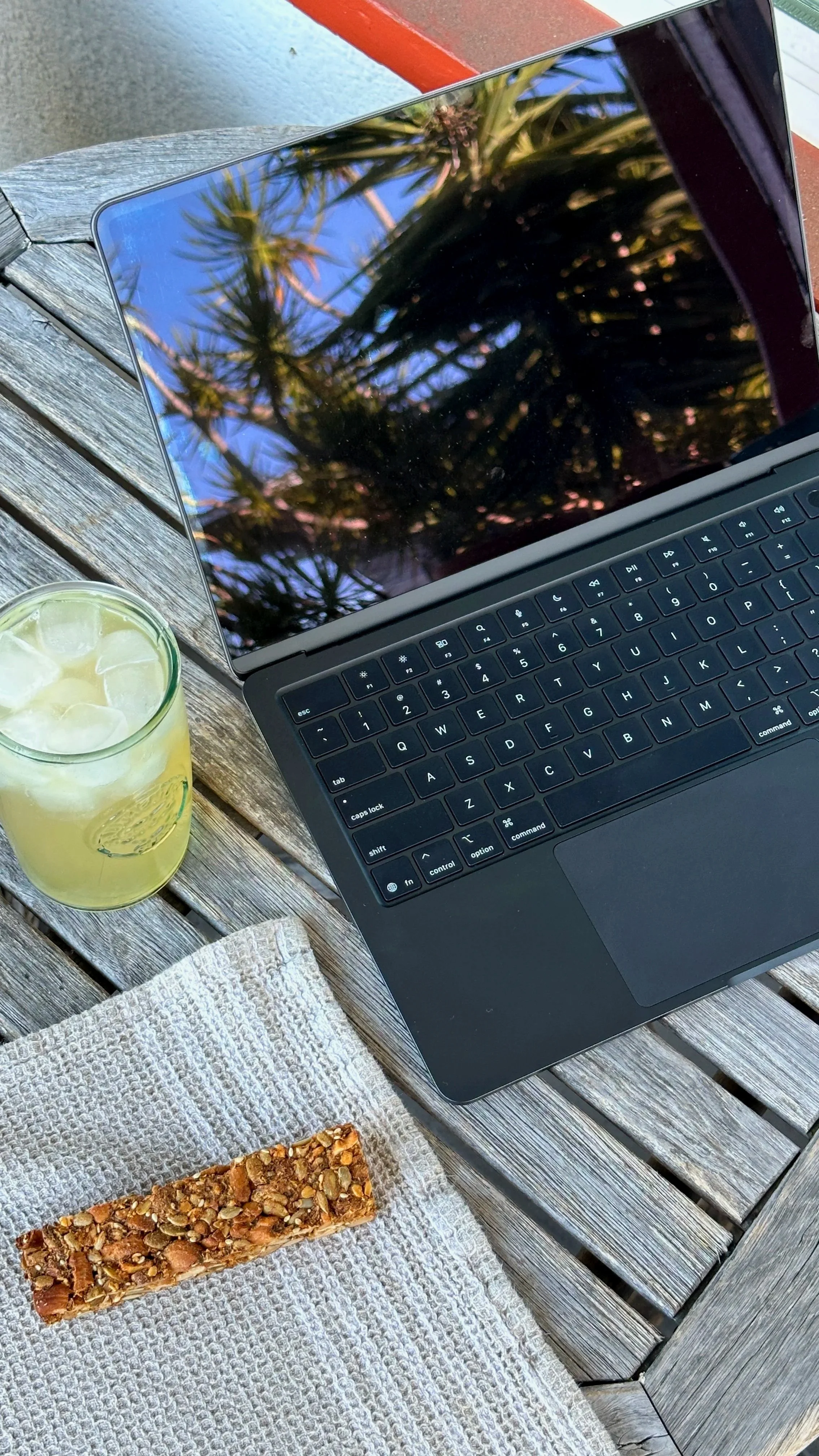 A laptop, a glass of lemonade with ice and lemon slices, and a granola bar on a white textured cloth on a weathered wooden outdoor table.