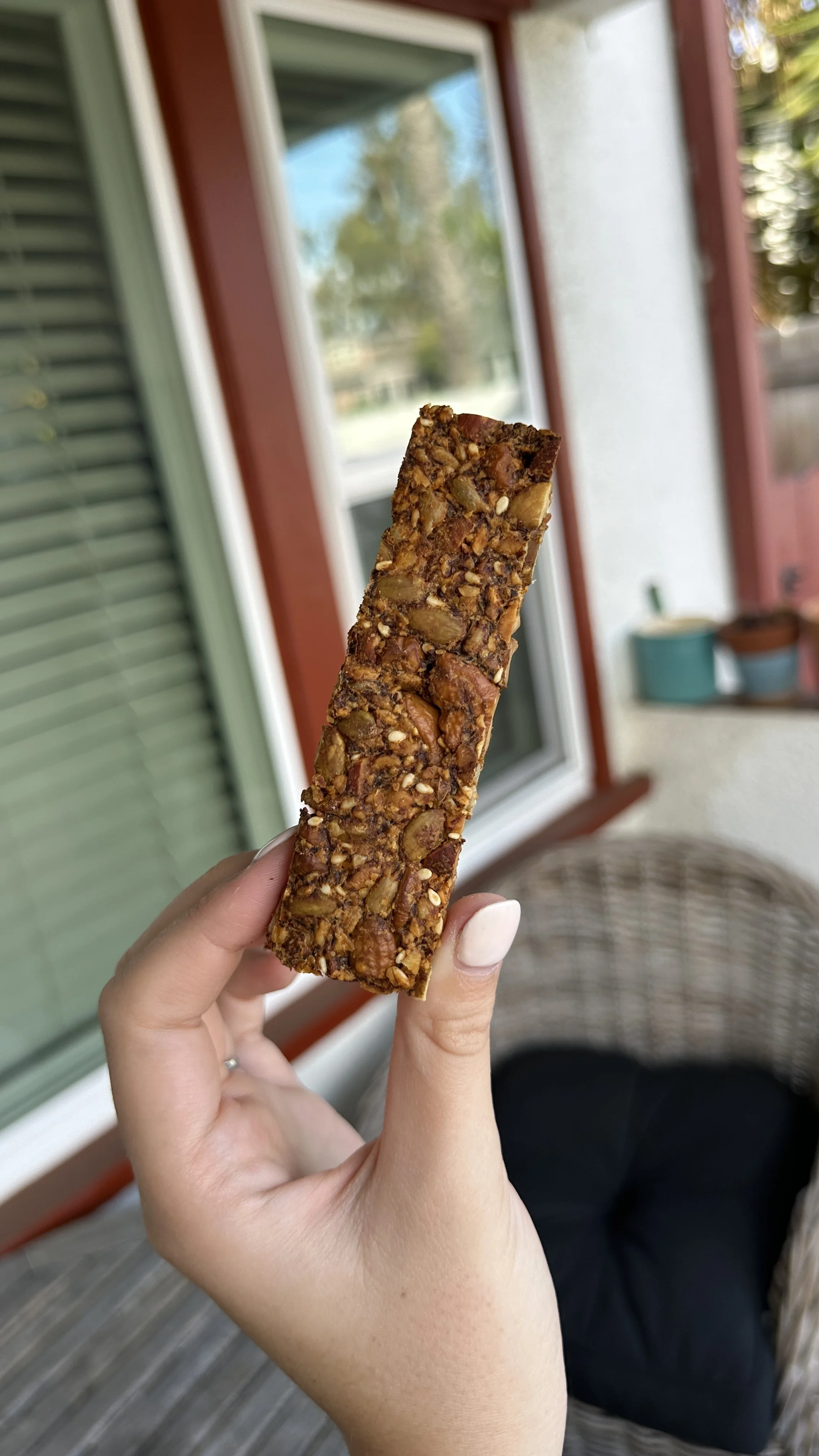 A hand holding a rectangular, nut-filled snack bar outside on a porch with window and patio furniture in the background.