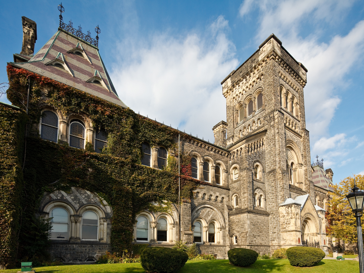 Historic stone castle with towers, arched windows, and ivy-covered walls, under a blue sky with clouds, surrounded by manicured bushes and grass.