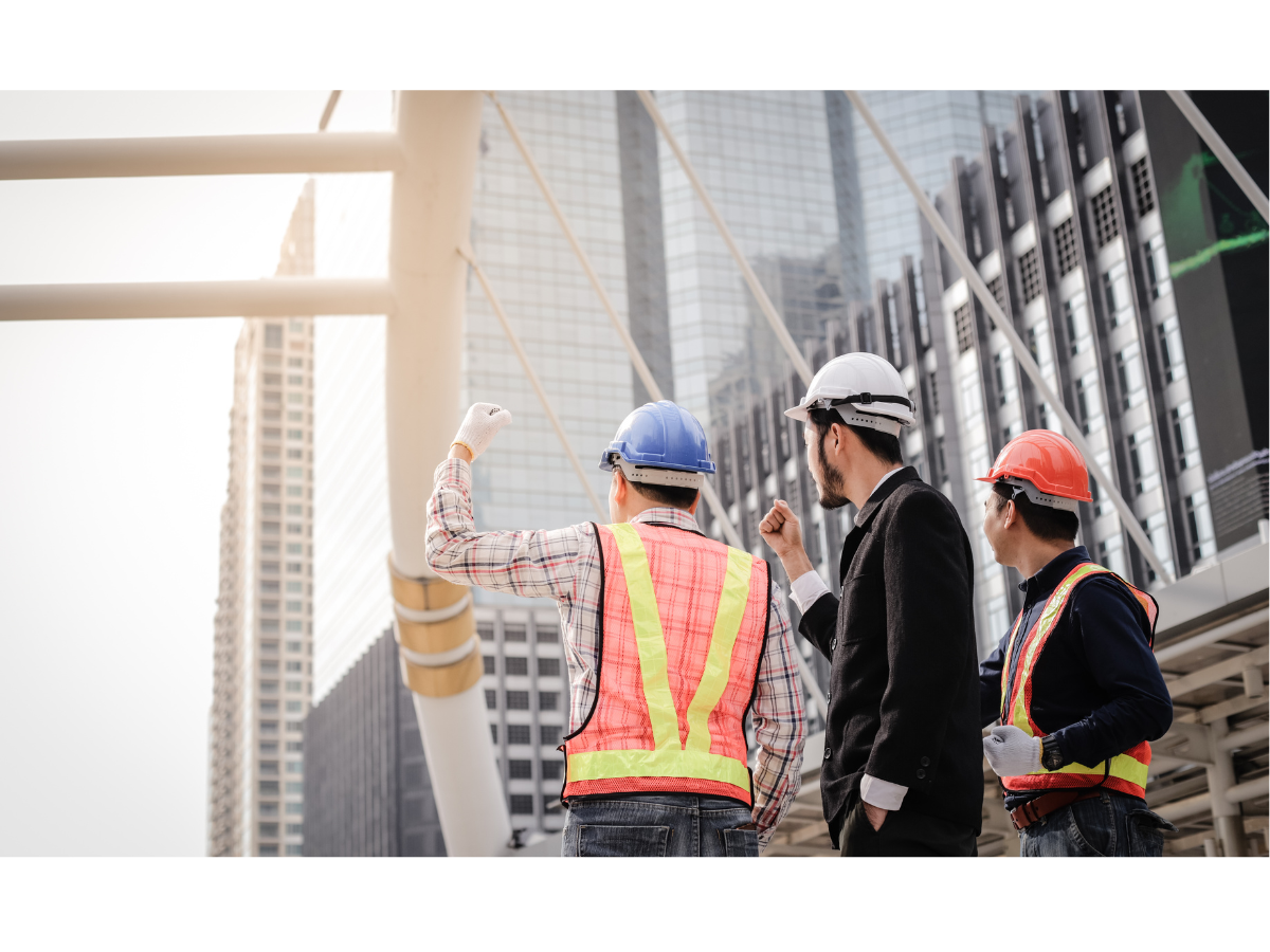 Three construction workers wearing safety helmets and vests, discussing at a construction site with tall buildings in the background.