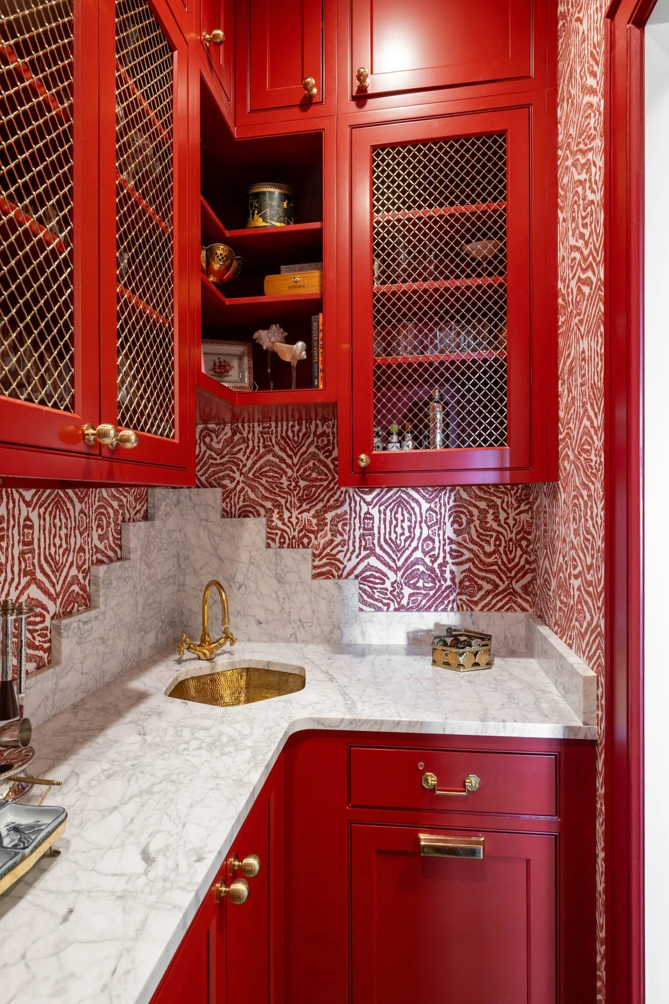 A small kitchen with red cabinets, gold hardware, a marble countertop and backsplash, and a gold sink faucet.