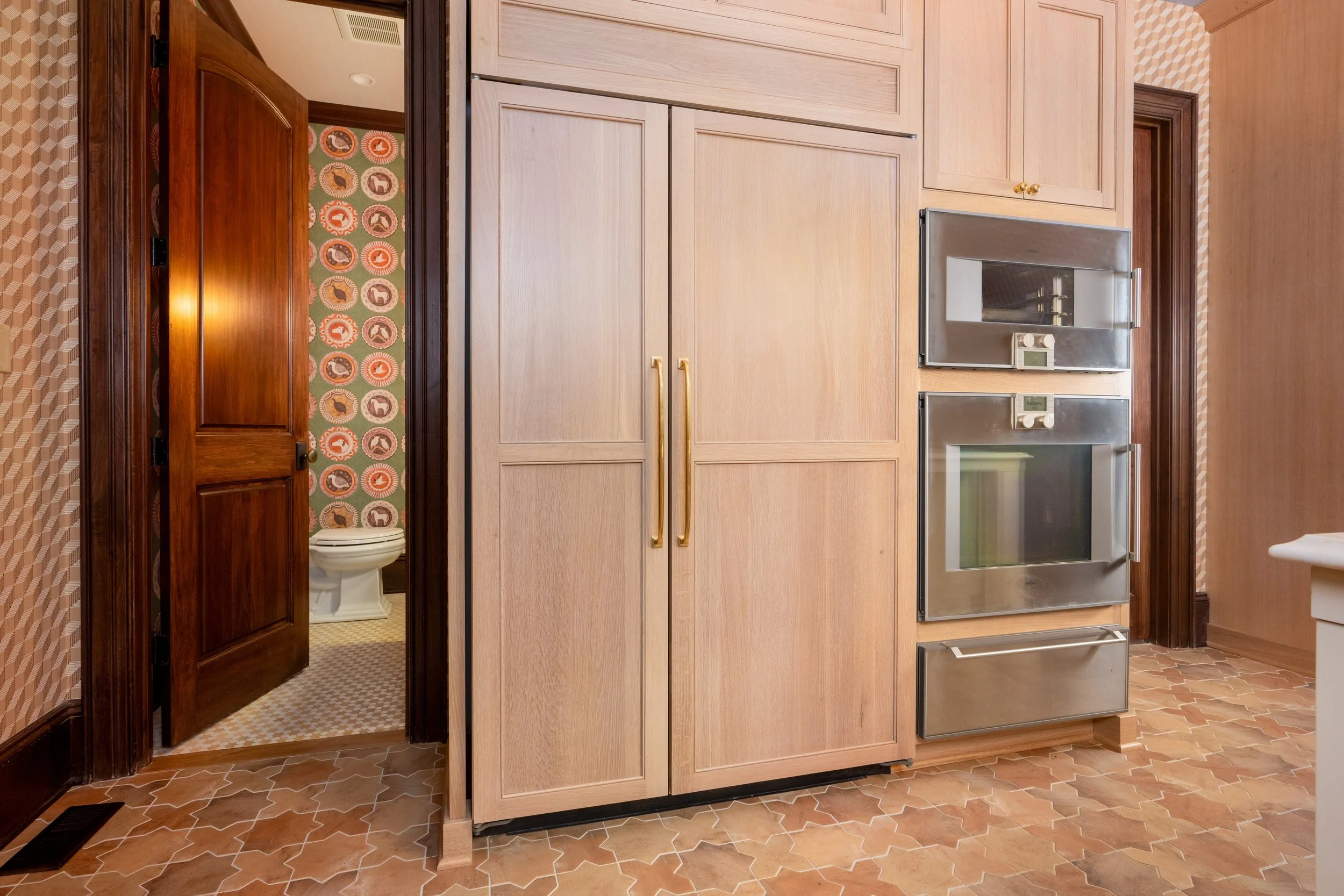Kitchen with light wood cabinetry and built-in stainless steel oven, and a view of a bathroom with a toilet and patterned wallpaper through an open door.