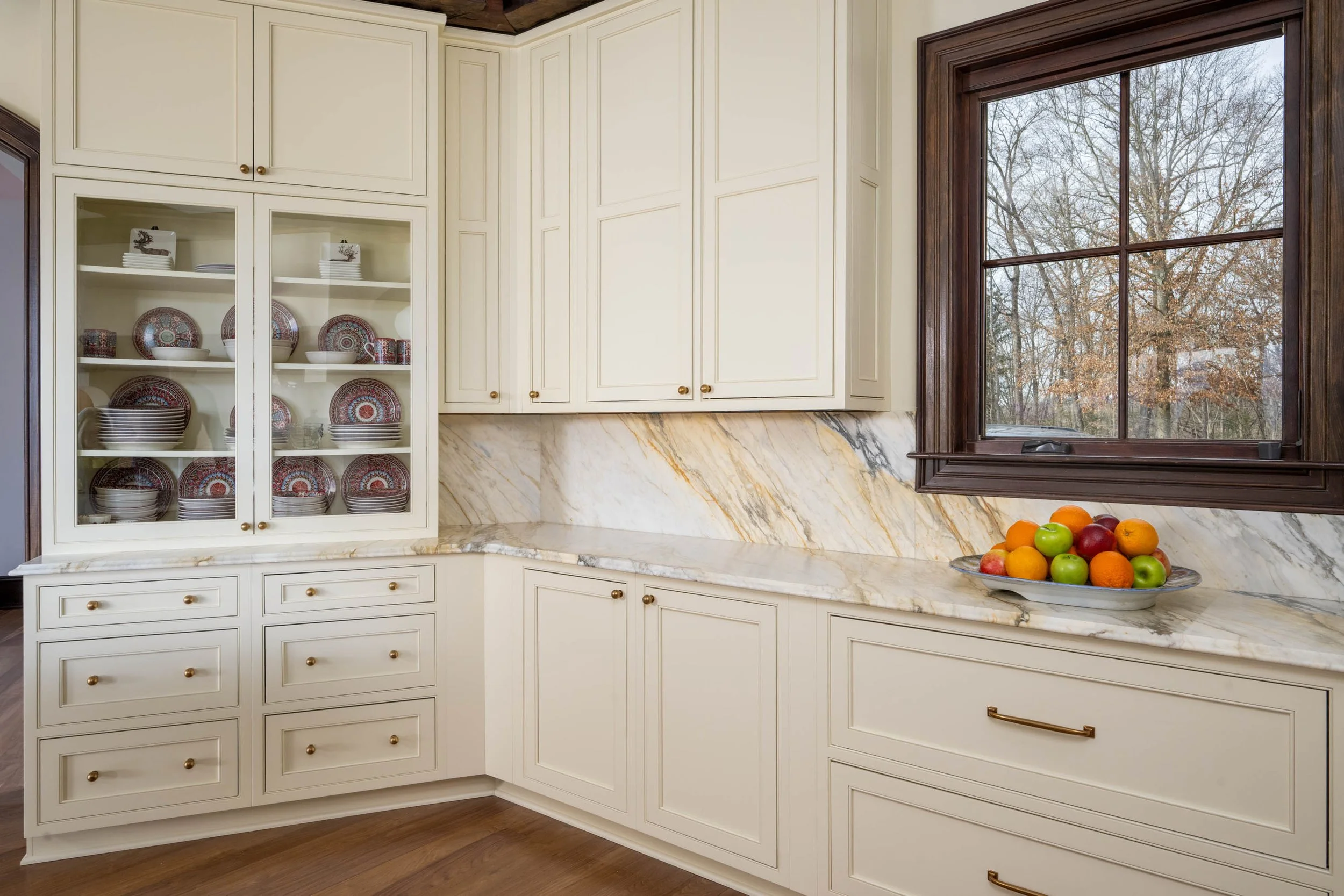 Kitchen corner with white cabinets, marble countertop and backsplash, a window with a view of trees, and a bowl of mixed fruit on the counter.