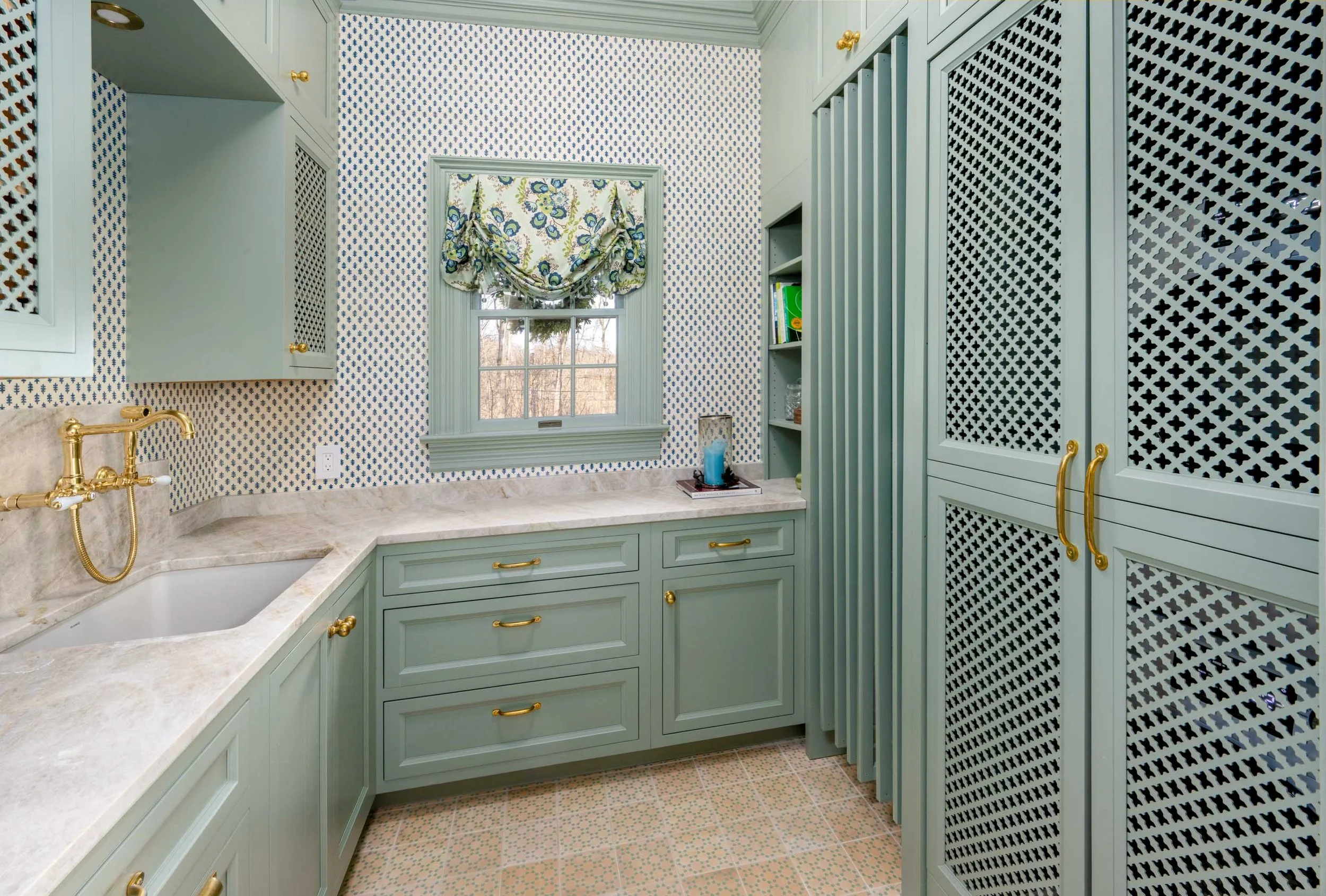 A vintage-style kitchen with pale green cabinets and gold hardware, a marble countertop, and a small window with a floral valance, decorated with patterned wallpaper and a shelving unit.