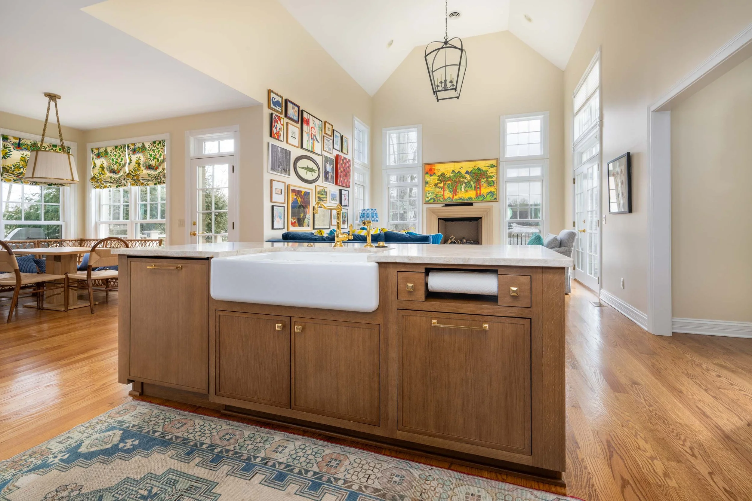 Open-concept living room and kitchen with hardwood floors, colorful art on the walls, large windows, custom cabinets, and a fireplace.