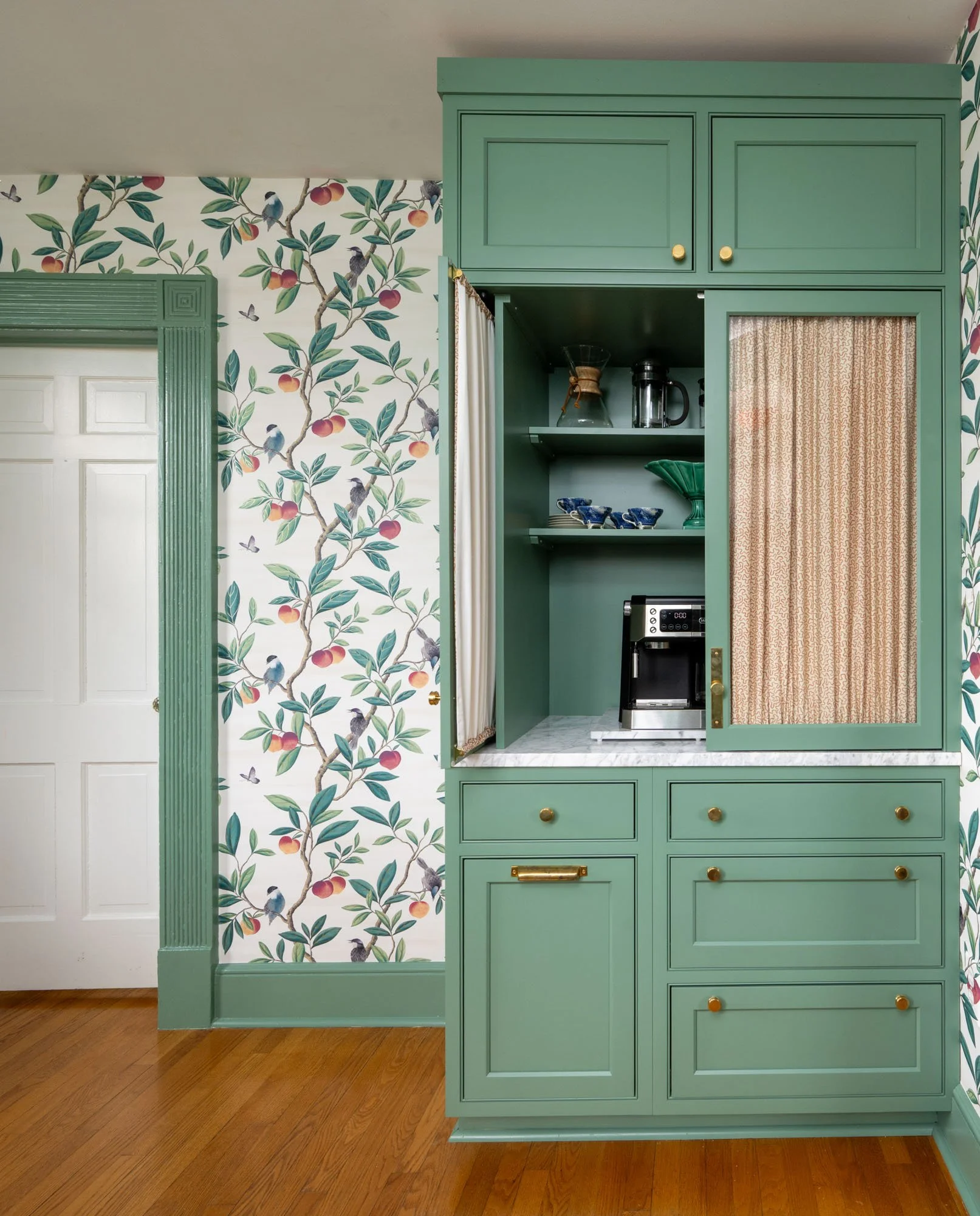 Custom coffee bar cabinet with green paint and gold hardware, with open shelves and a coffee machine, next to floral wallpaper with birds and fruit, and a white door.