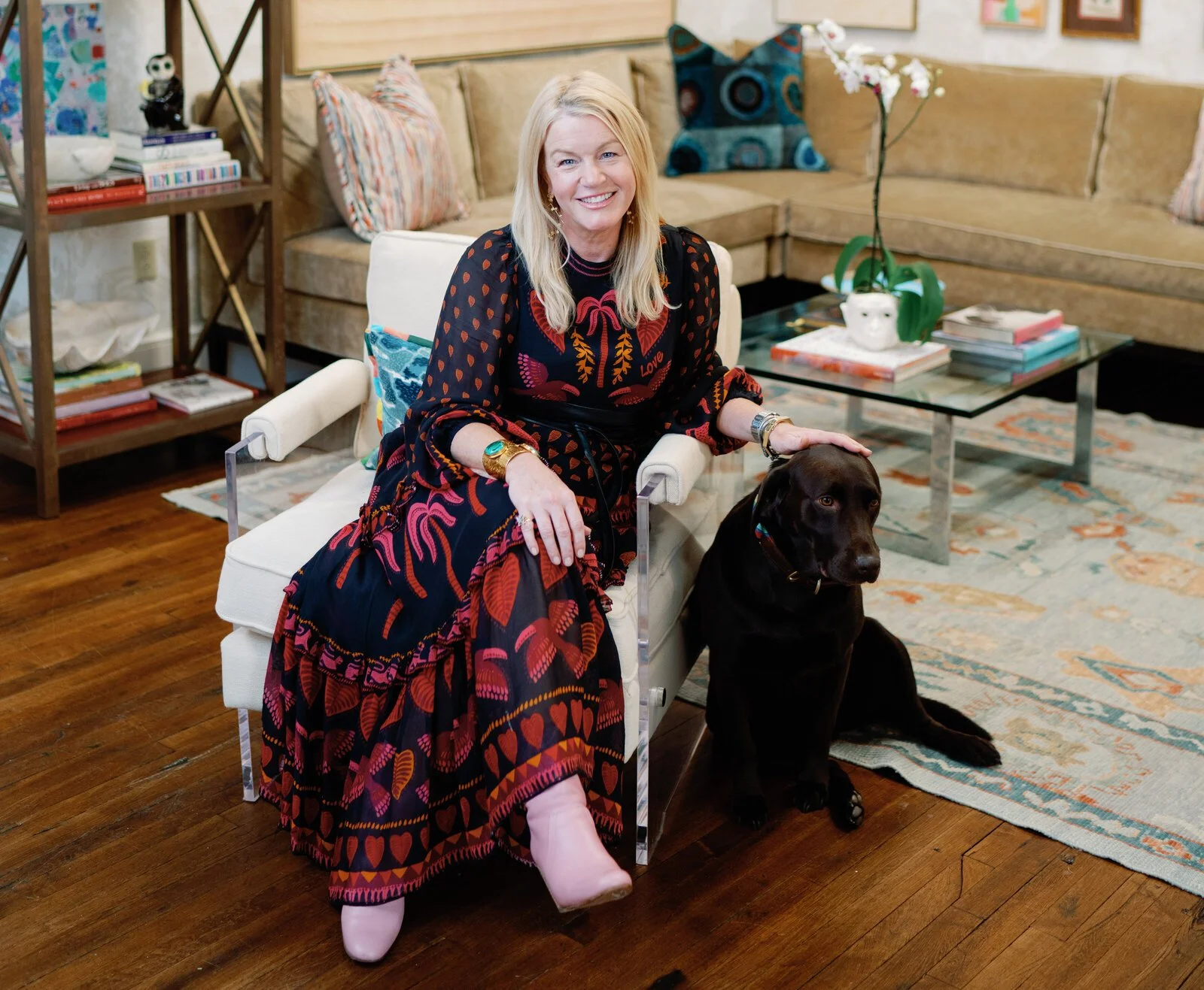 A woman, interior designer and Guild Cabinetry owner Mary Caroline Mumpower, in a colorful dress sitting on a white chair with a black Labrador retriever in a living room.