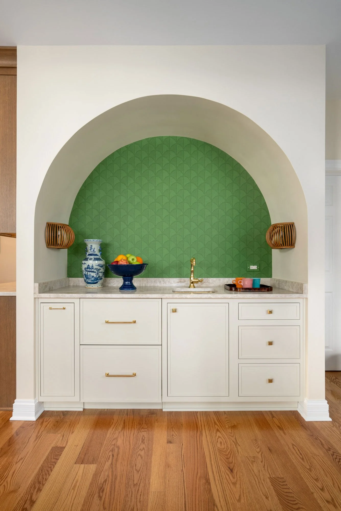 Coffee bar area with a green arched alcove, white cabinets with gold hardware, and a marble countertop. Decor includes a blue and white vase, a bowl of fruit, and colorful cups. Wooden wall sconces are on either side of the alcove.