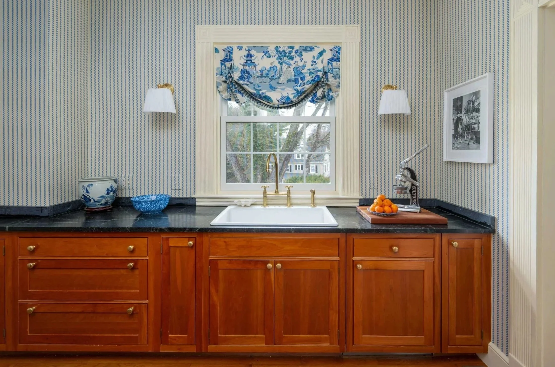 Kitchen sink with window above, blue and white patterned curtains, wooden custom cabinetry, black countertop, brass faucet, small decorative items, framed picture, and wall-mounted lamps.