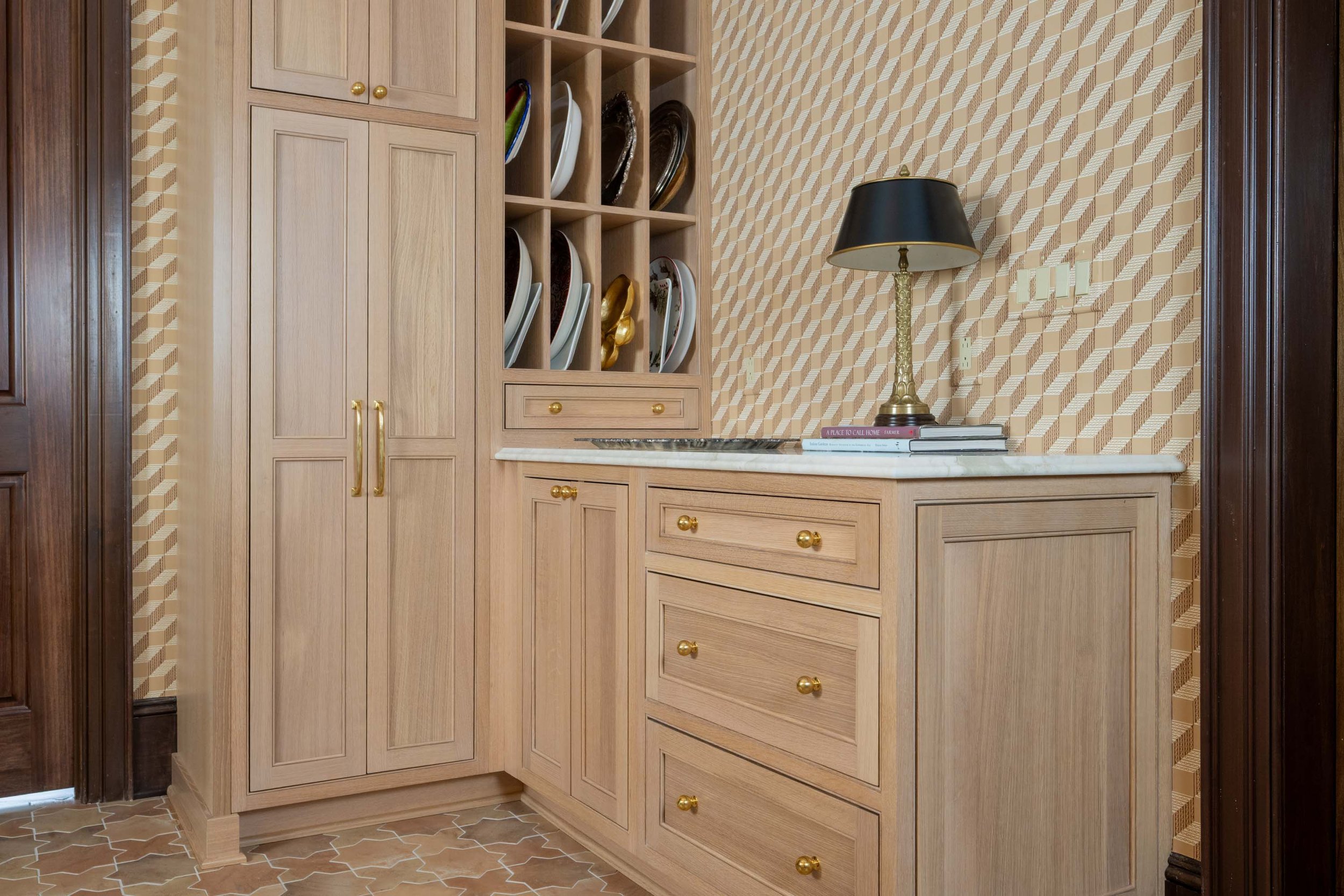 A kitchen corner with wooden cabinets, open shelves with plates, a lamp, and books on a marble countertop.