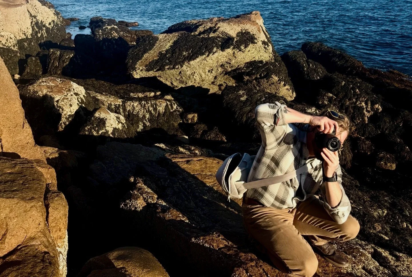 Person crouching on rocks near the water, holding a camera up to their face, with a backpack and sunglasses, during sunset or sunrise.