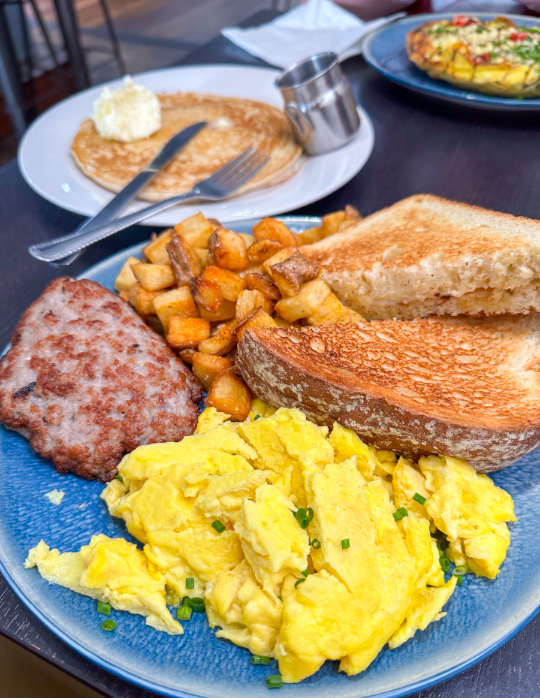 Scrambled eggs, sausage, toast, and potatoes on a blue plate. Pancakes with butter and syrup nearby. Bright, appetizing breakfast scene.