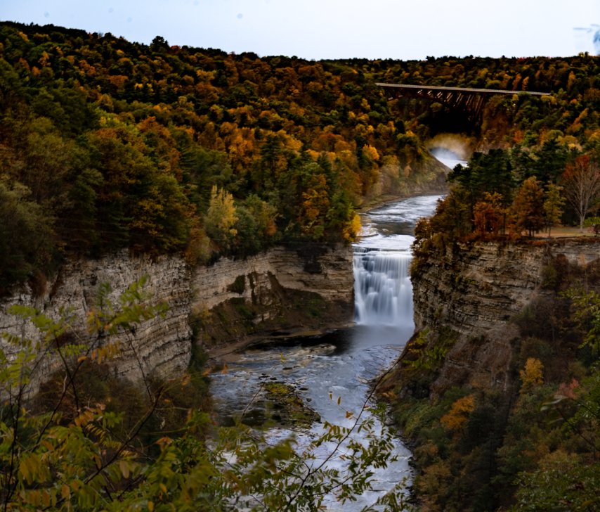 Waterfall cascading between rocky cliffs in an autumn forest, with vibrant fall foliage. A bridge spans the gorge in the background.