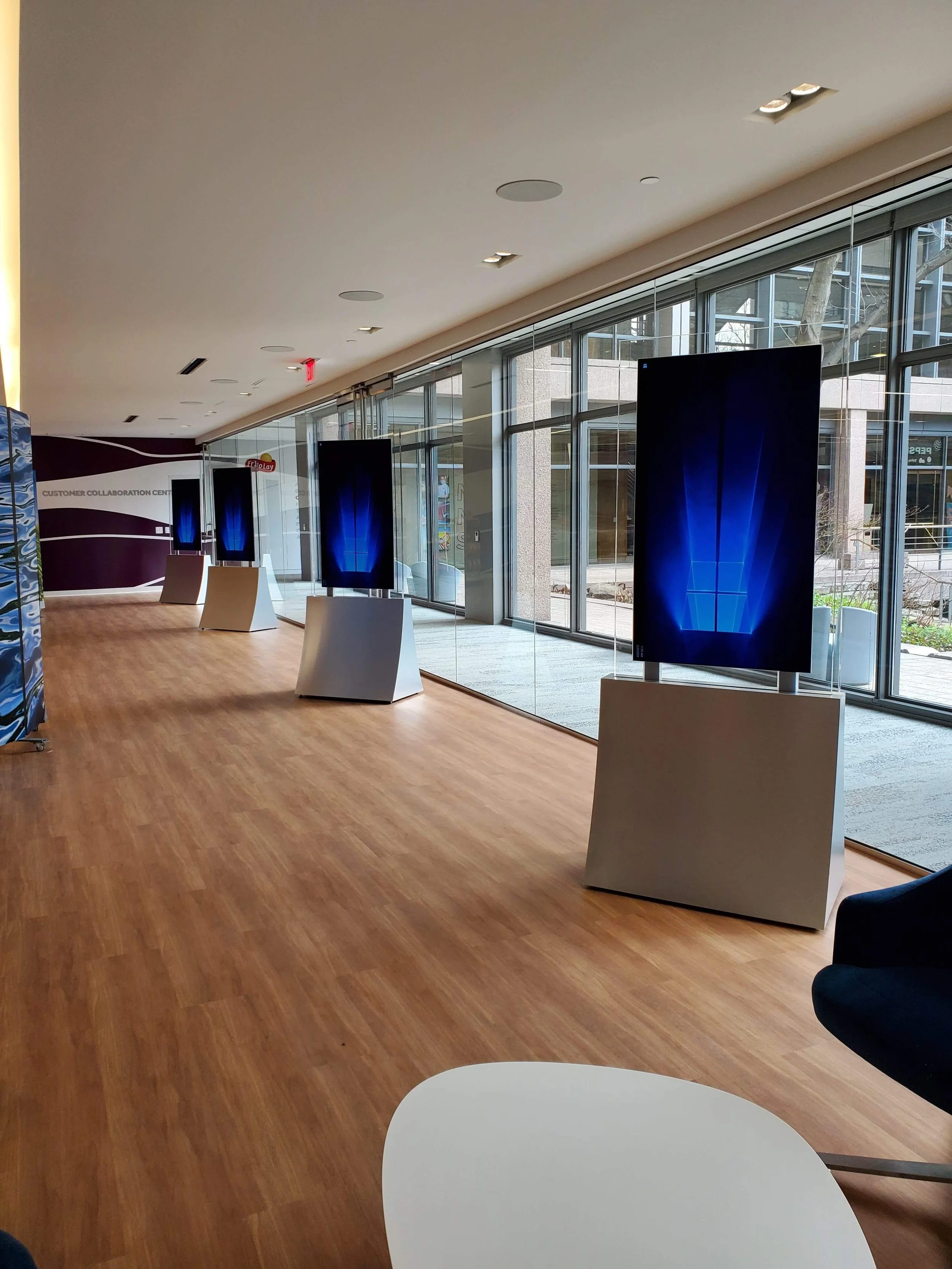 Interior of a modern technology demonstration area with large screens mounted on white stands, a glass wall facing outside, and a wooden floor.