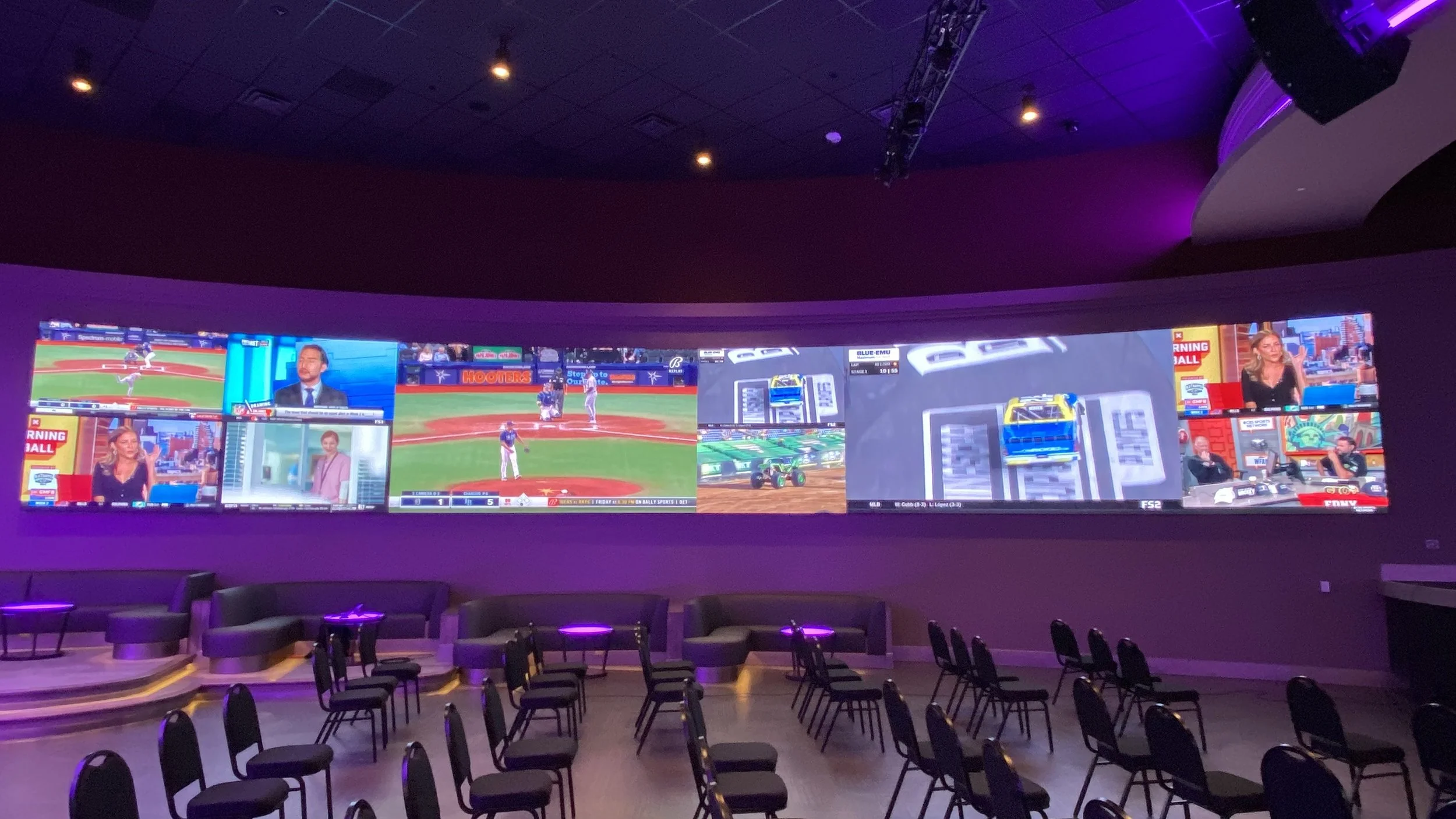 Multiple television screens displaying sports and news channels in a dark room with purple lighting and chairs arranged in front.
