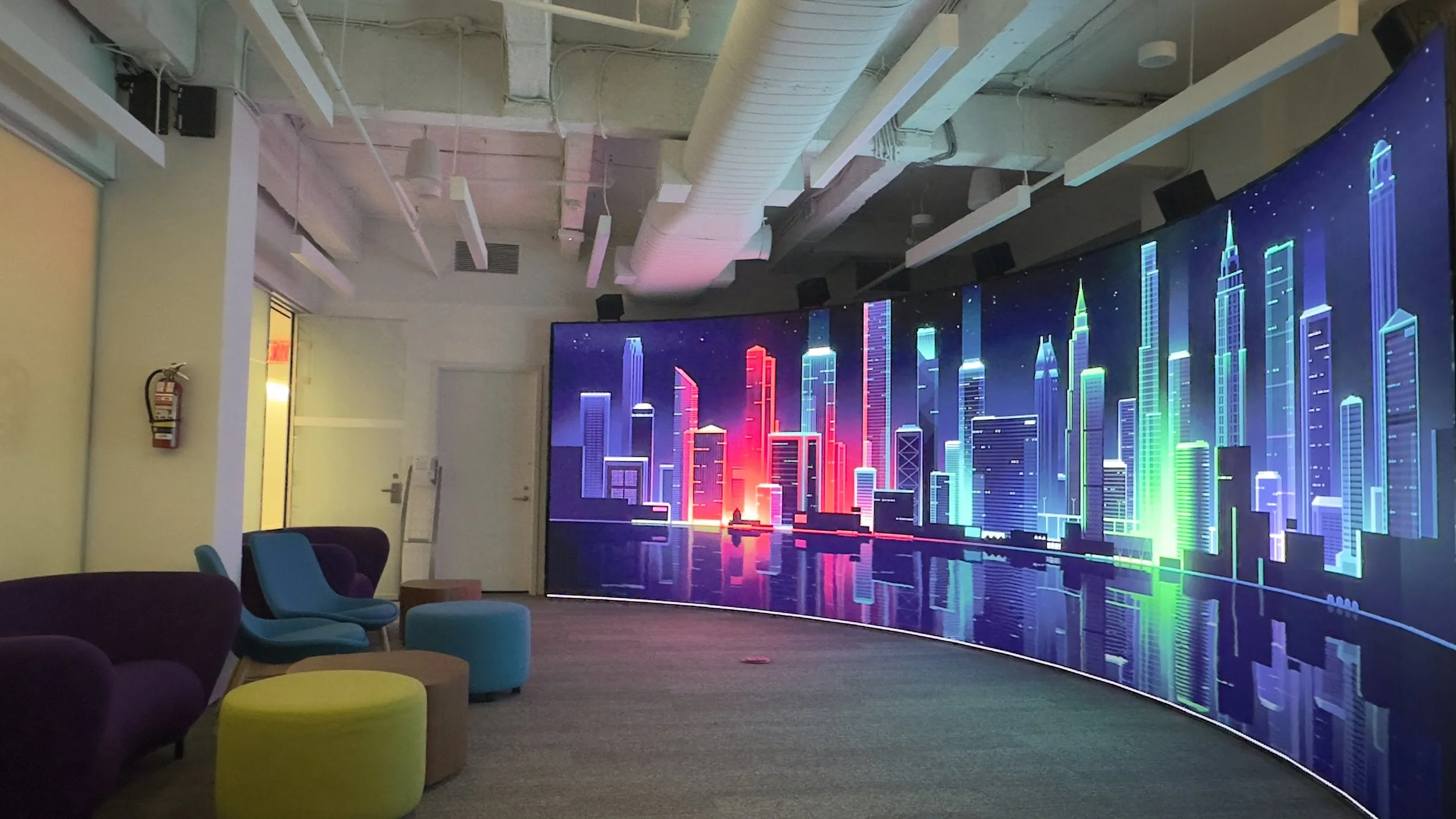 Interior room with colorful LED cityscape display, including chair and ottoman seating, fire extinguisher on wall, and ceiling pipes and vents.
