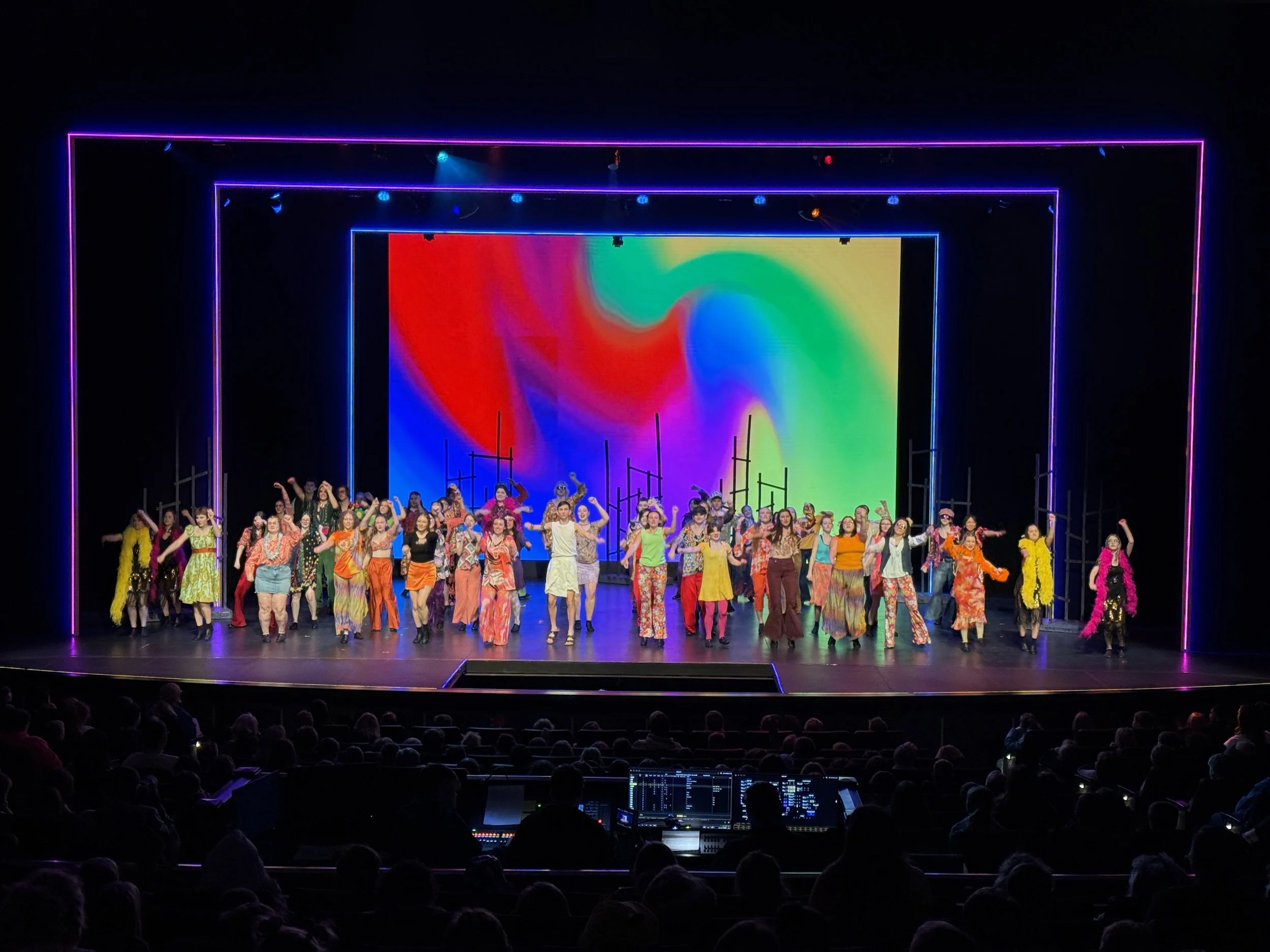 Group of performers dancing on stage with colorful background and neon frame.