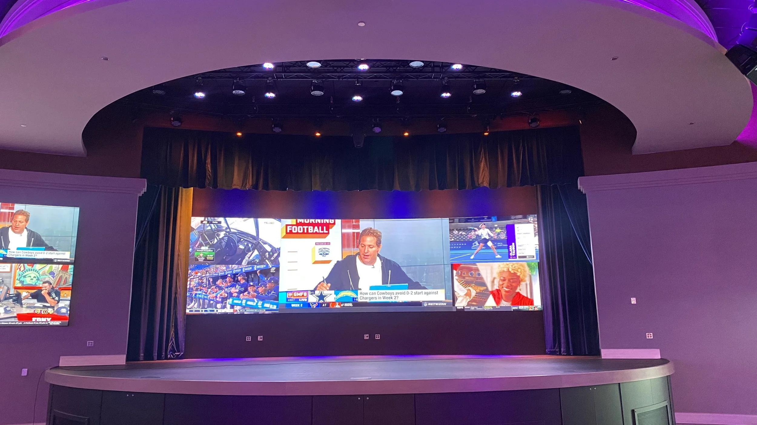 An indoor stage with a large wall of multiple screens displaying sports coverage, including football and tennis, with a person speaking at the center.