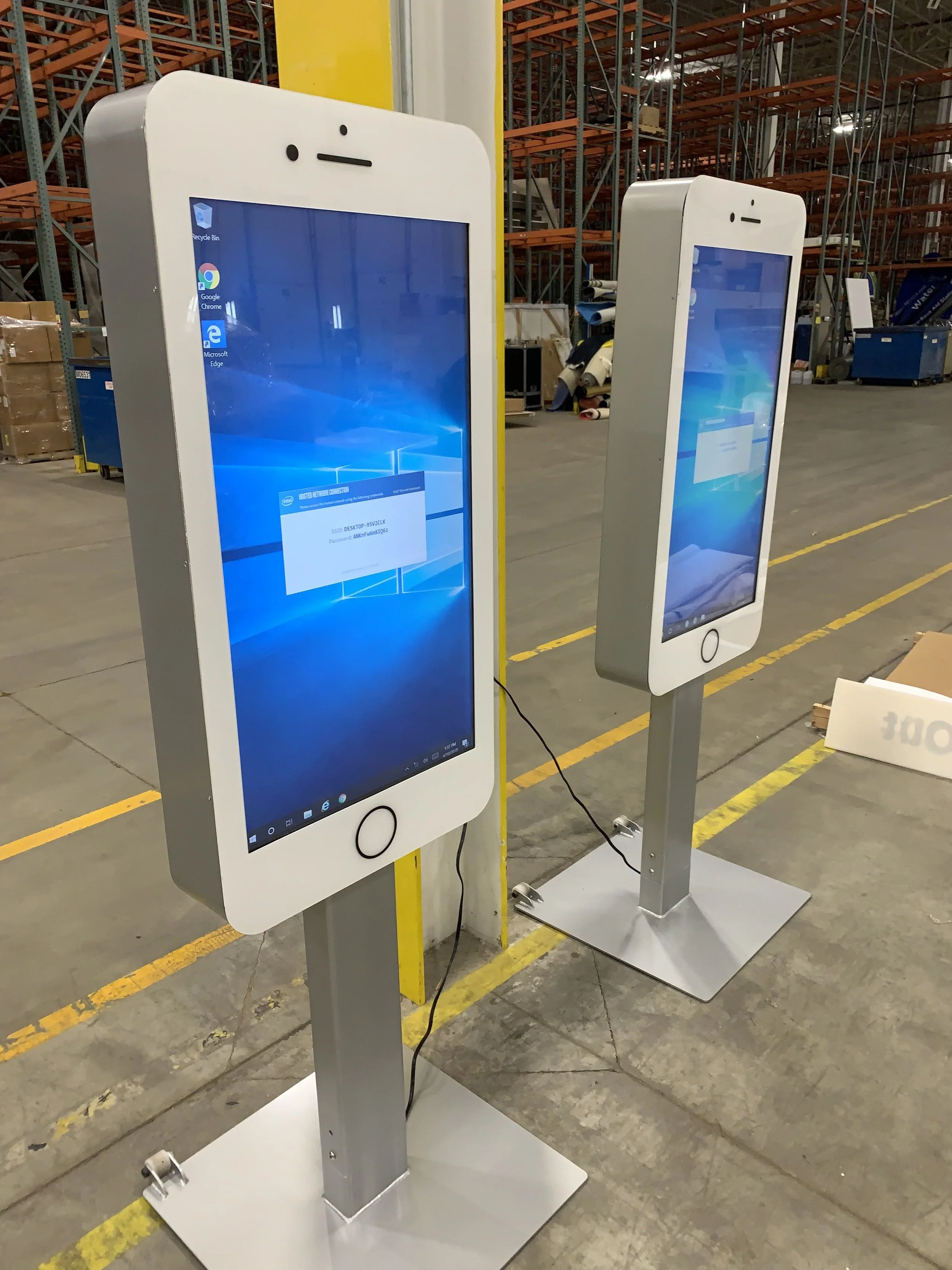 Two large touchscreen kiosks shaped like smartphones in an industrial warehouse setting.
