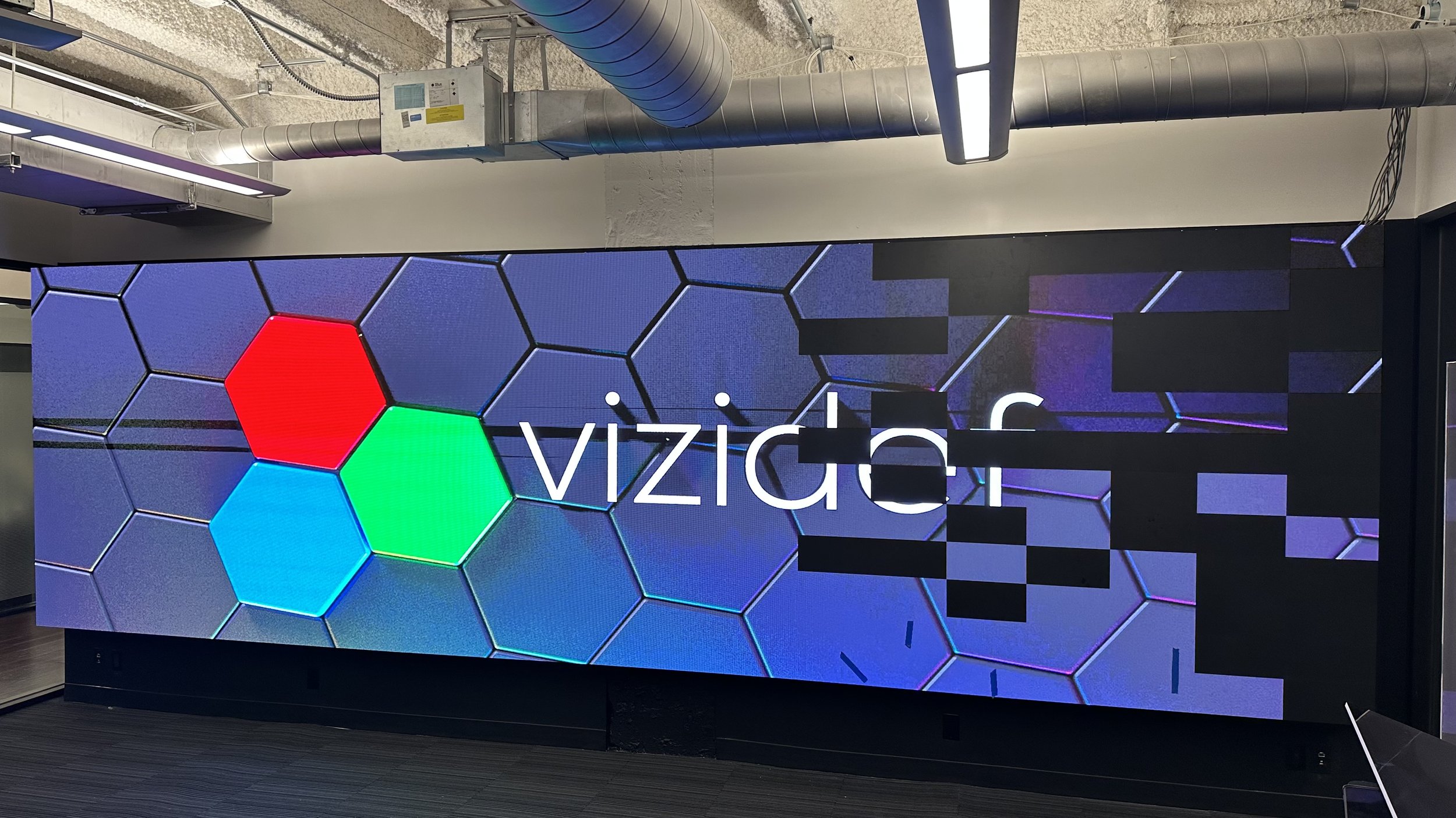 Digital screen displaying a logo with hexagonal shapes, two are red and blue and one is green, behind the partially obscured white text 'vizion'.