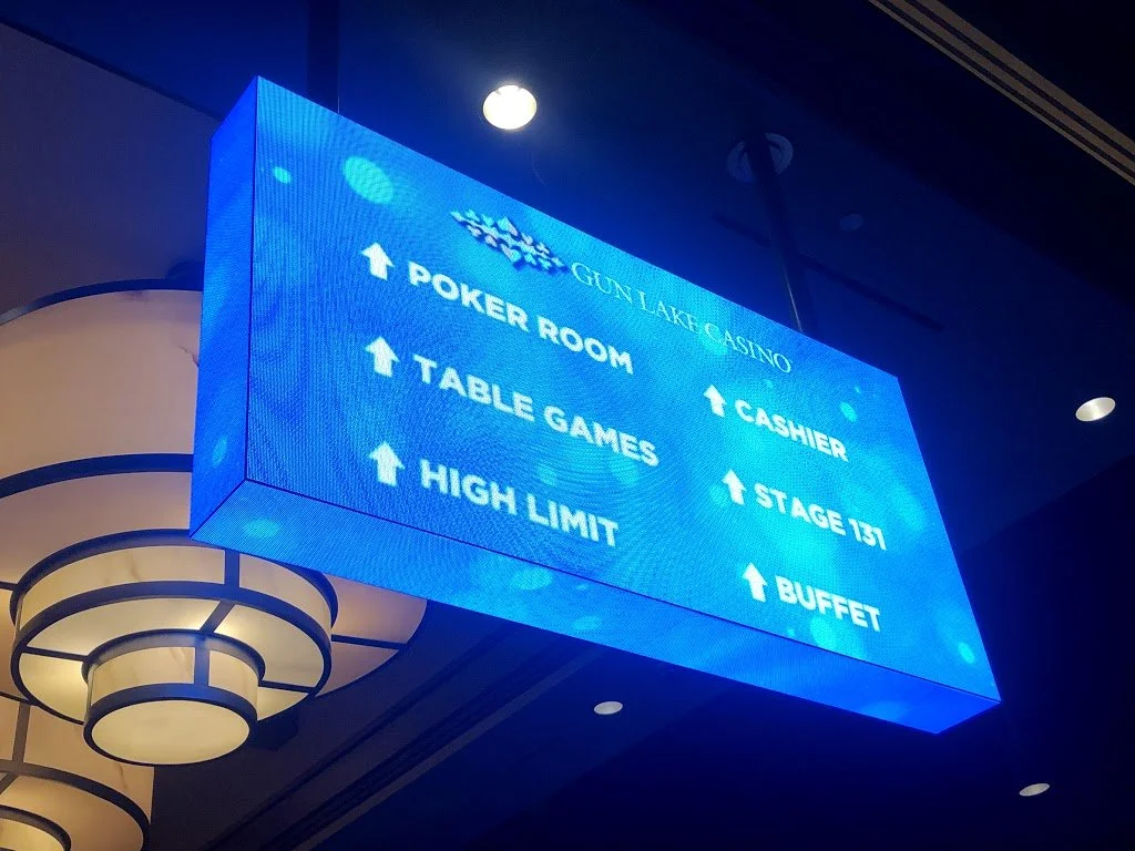 Blue digital sign at Gun Lake Casino listing directions to poker room, table games, high limit, cashier, stage 131, and buffet, with ceiling lights visible.
